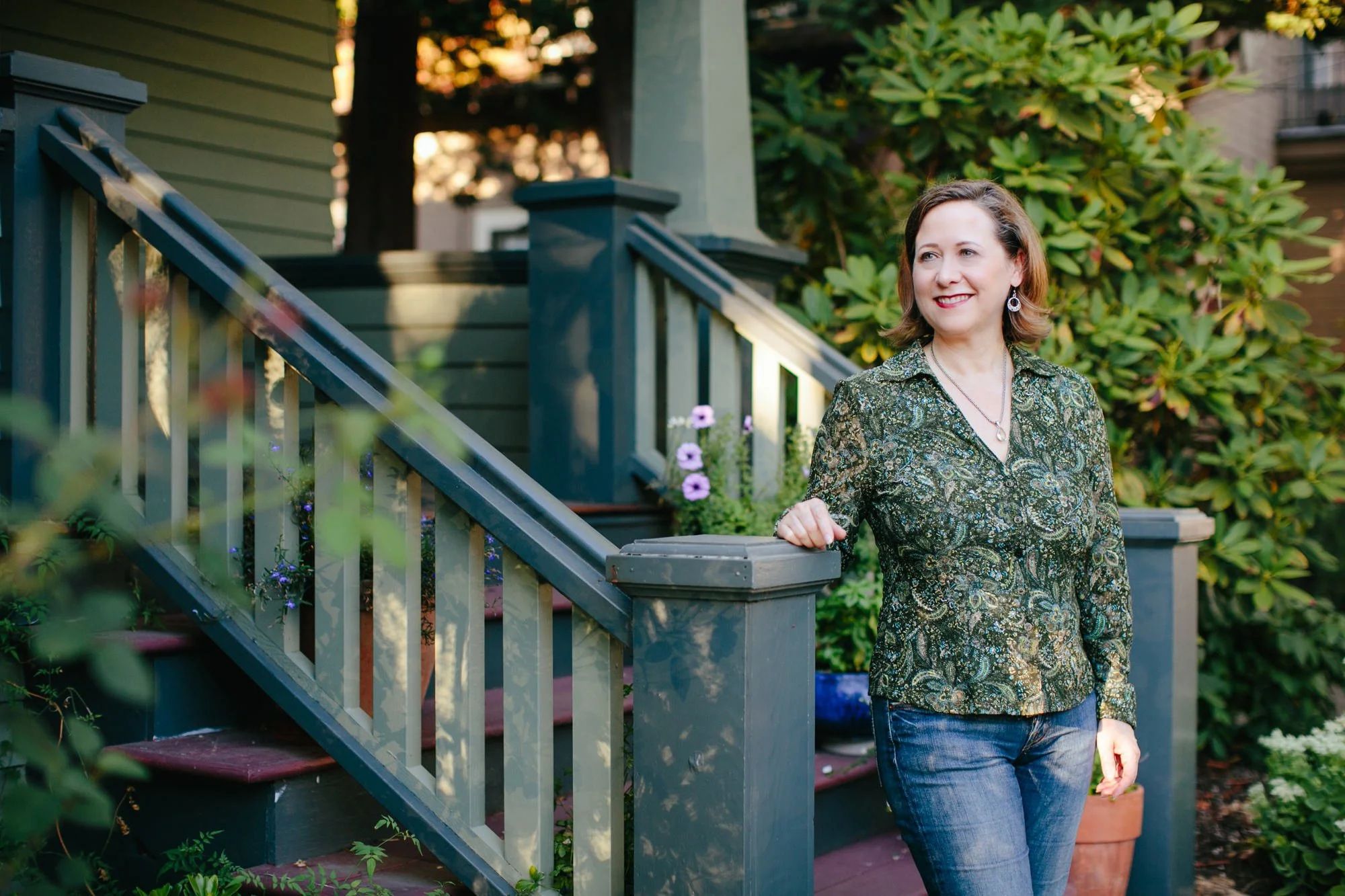 Woman smiling and standing on a porch with a garden behind her, wearing a patterned blouse and jeans.