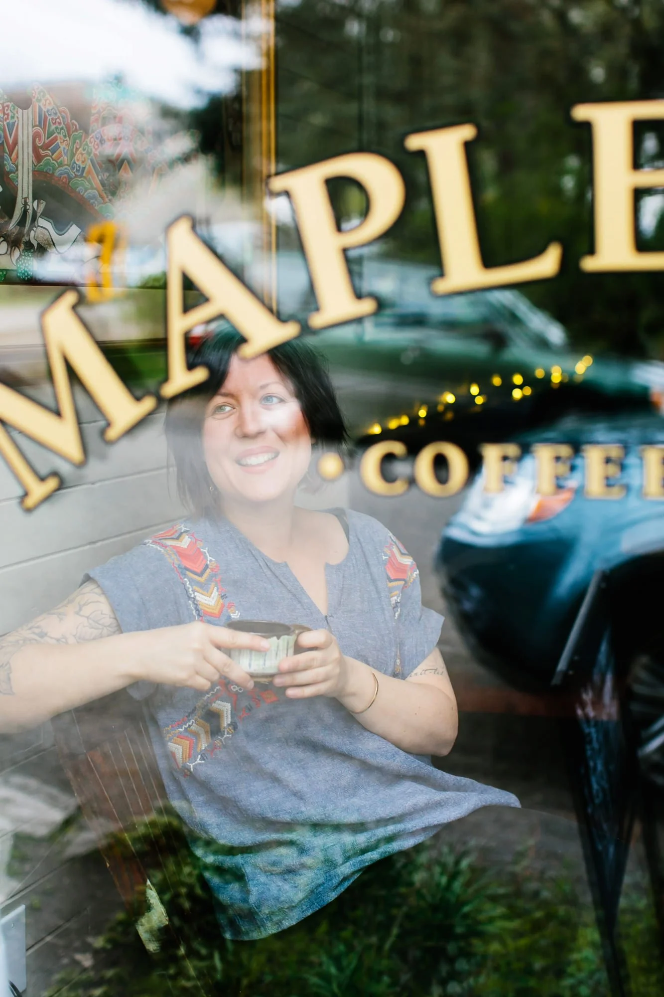 Woman with dark hair and tattoos sitting inside a coffee shop, smiling and holding a cup, seen through a glass window with the shop's name and coffee offerings on it.
