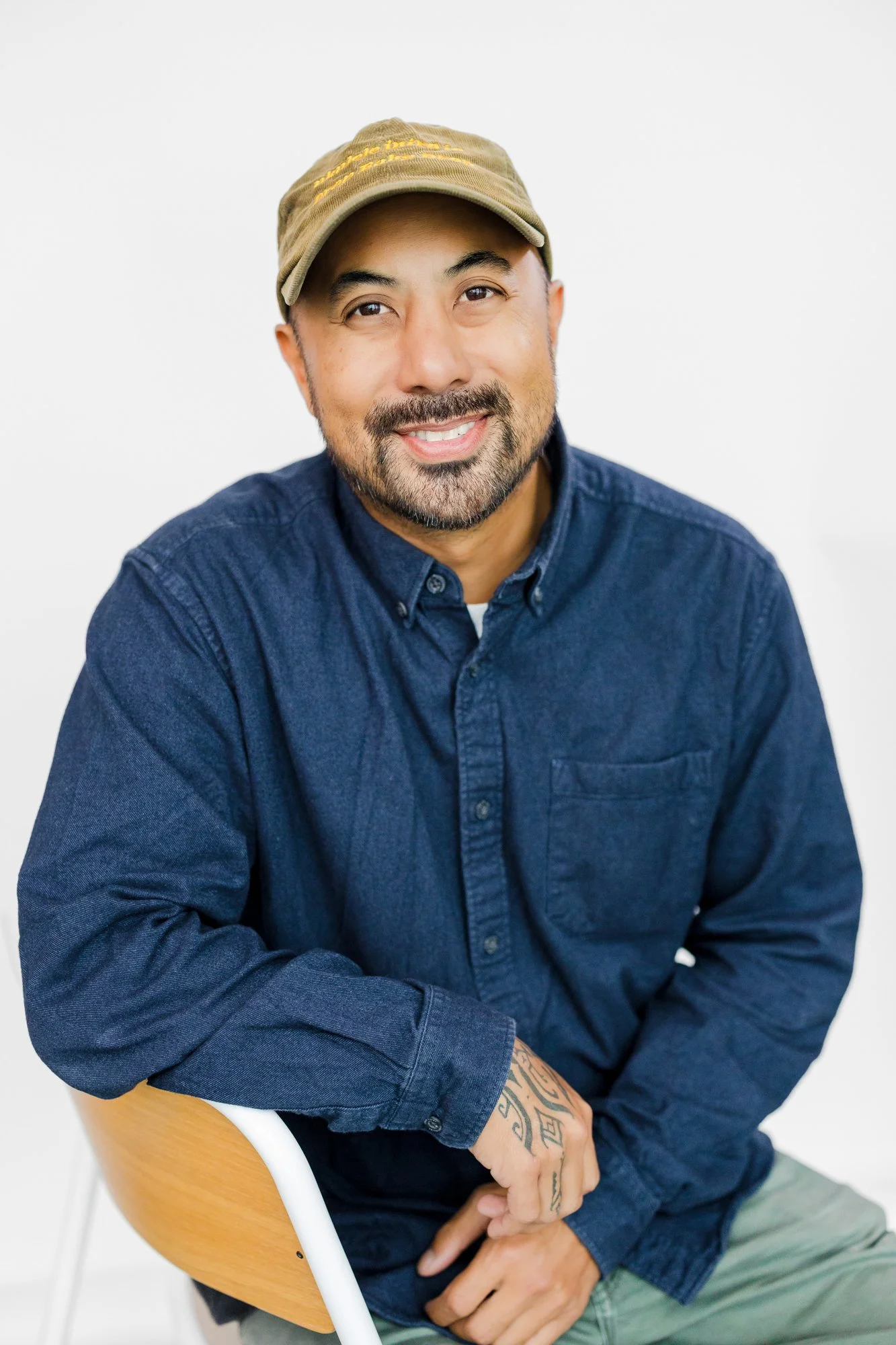 A man with a beard and mustache wearing a tan cap, navy blue button-up shirt, and khaki pants, sitting on a wooden chair with a white background.