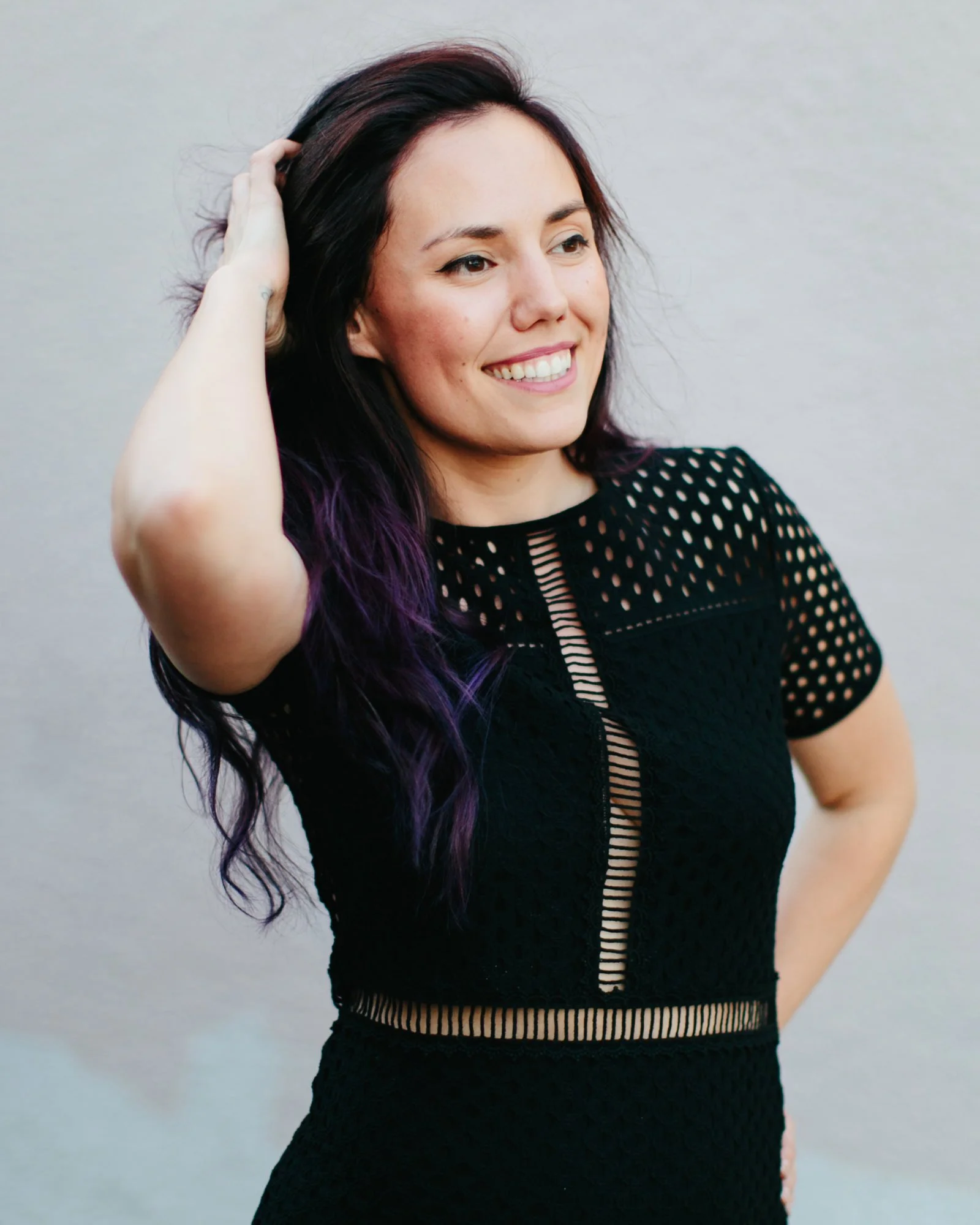 A smiling woman with dark hair and purple highlights, wearing a black perforated dress, standing against a plain light background.