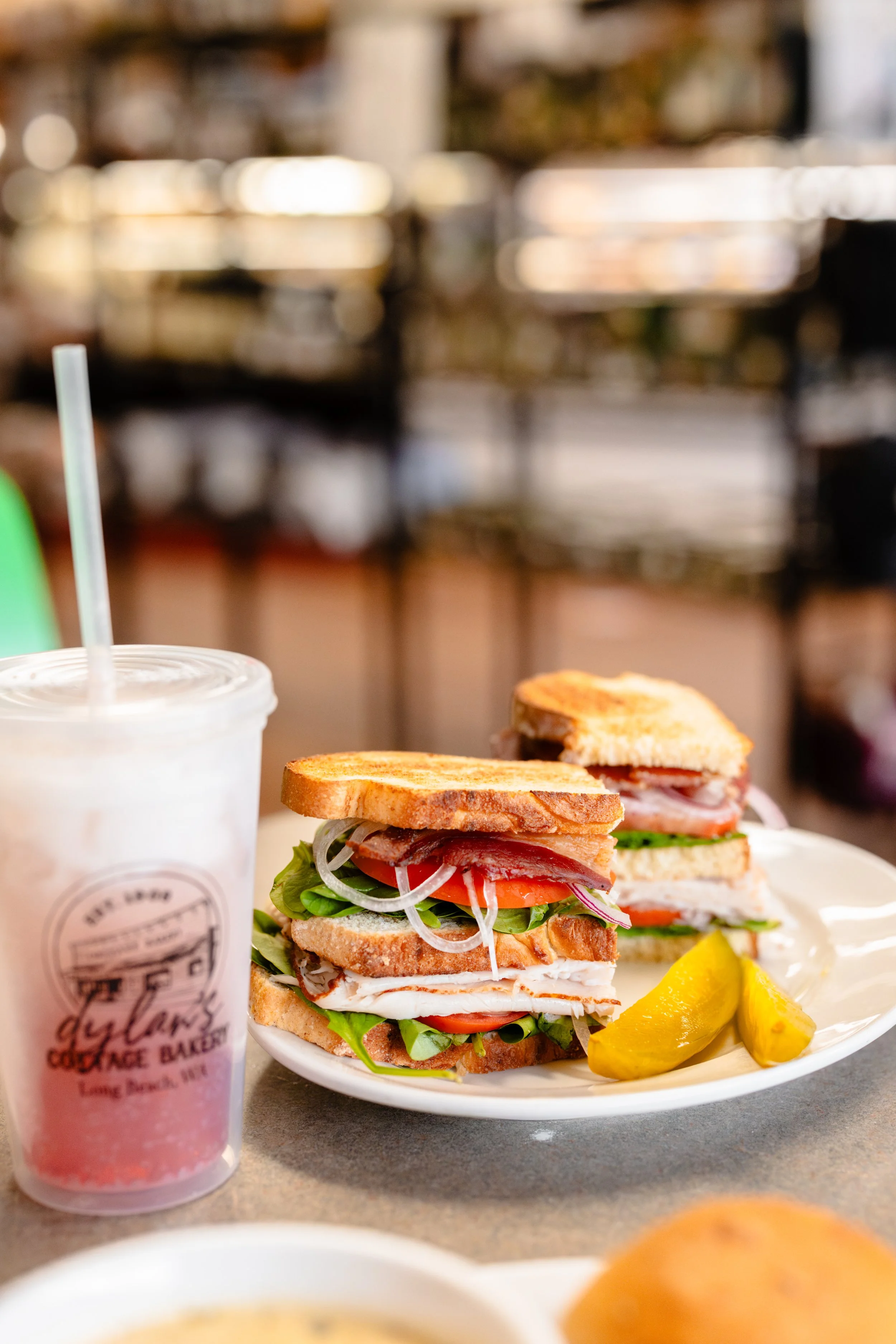 A sandwich with lettuce, tomato, onions, turkey, and bacon on toasted bread, served with pickles on a white plate, and a pink drink in a clear cup with a straw.