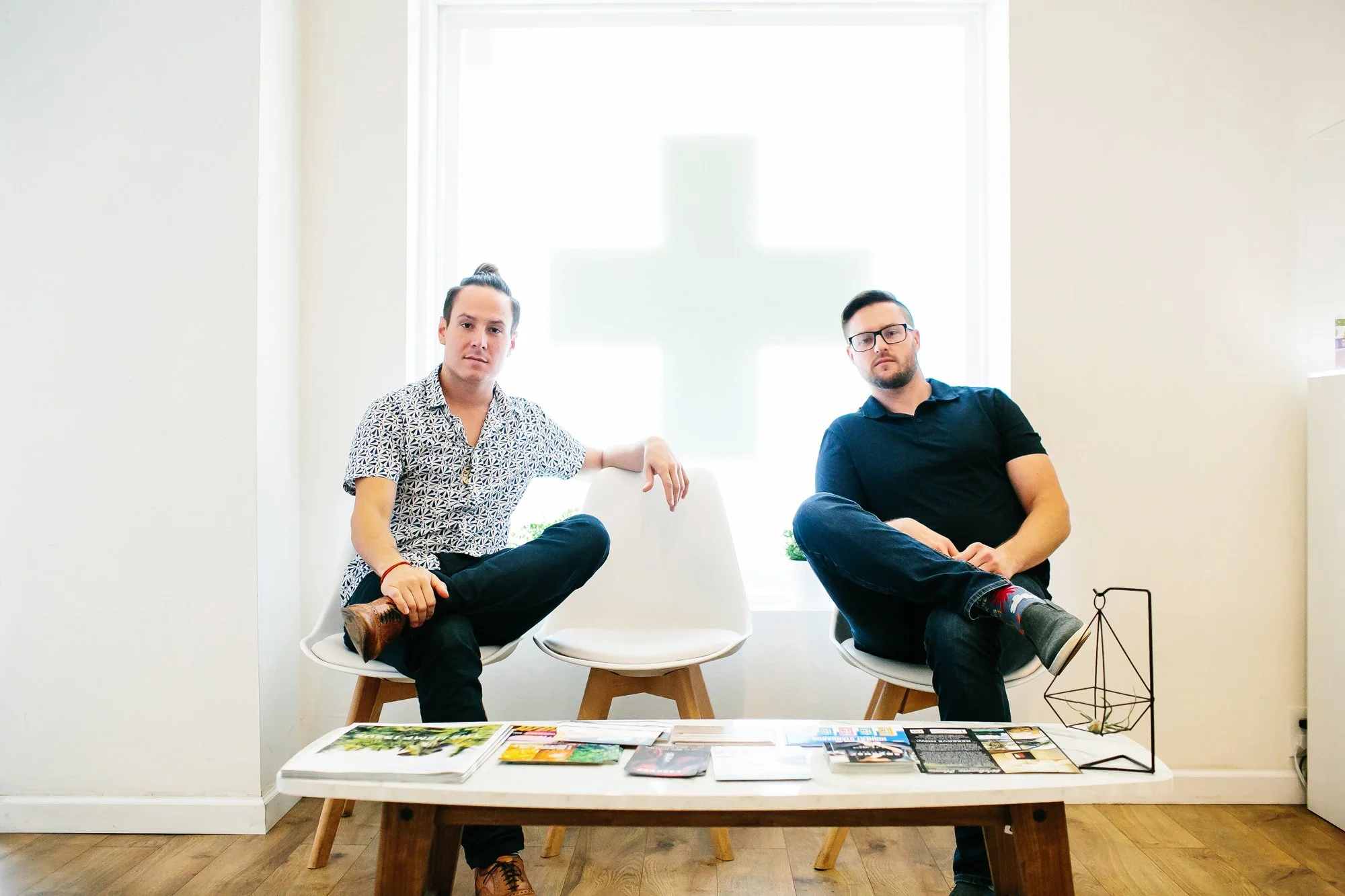 Two young men sitting on white chairs in a room with a cross-shaped window behind them. They are facing forward with magazines on a low table in front of them.