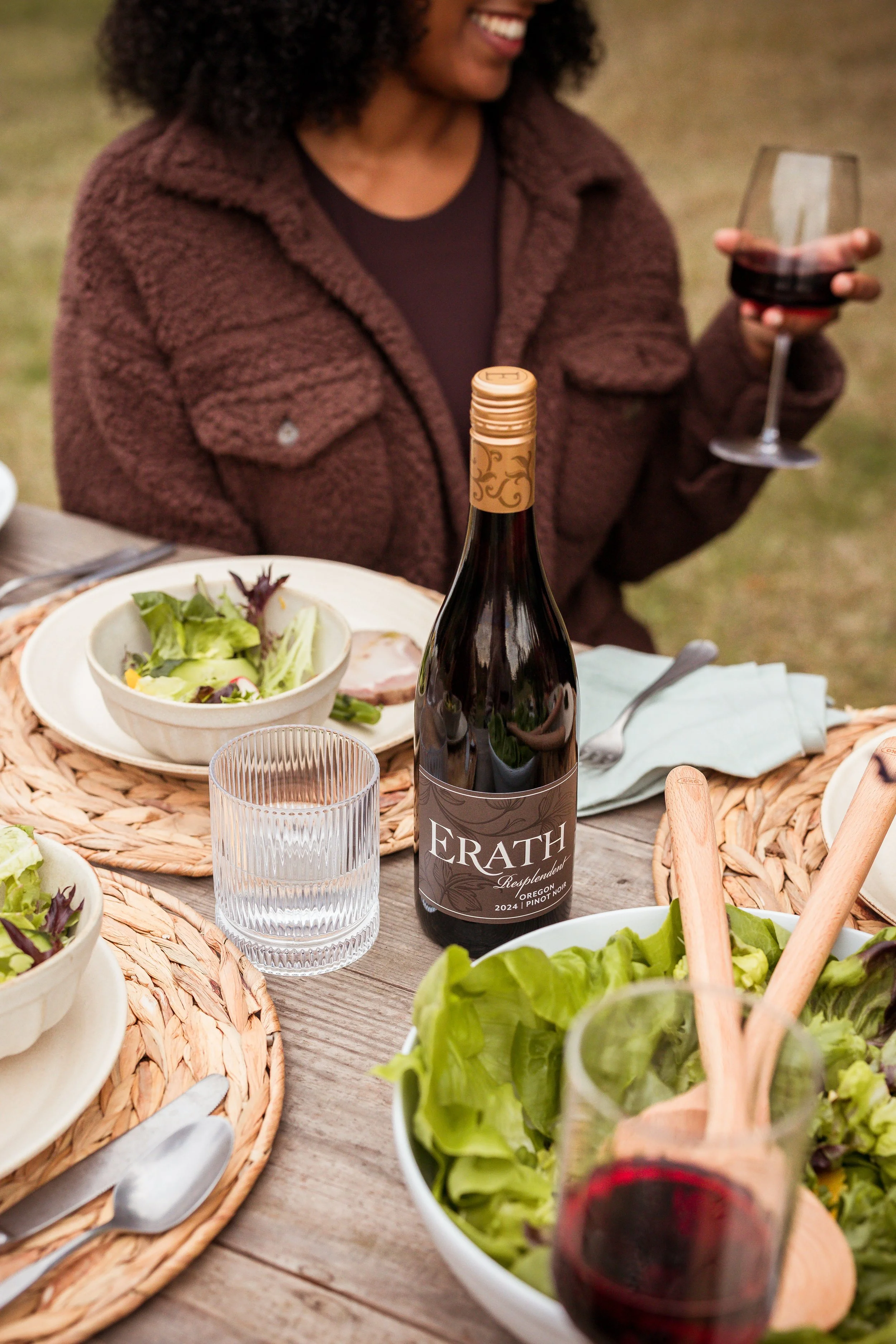 A woman holding a glass of red wine at an outdoor meal table with salads, a bottle of Erath wine, and utensils.