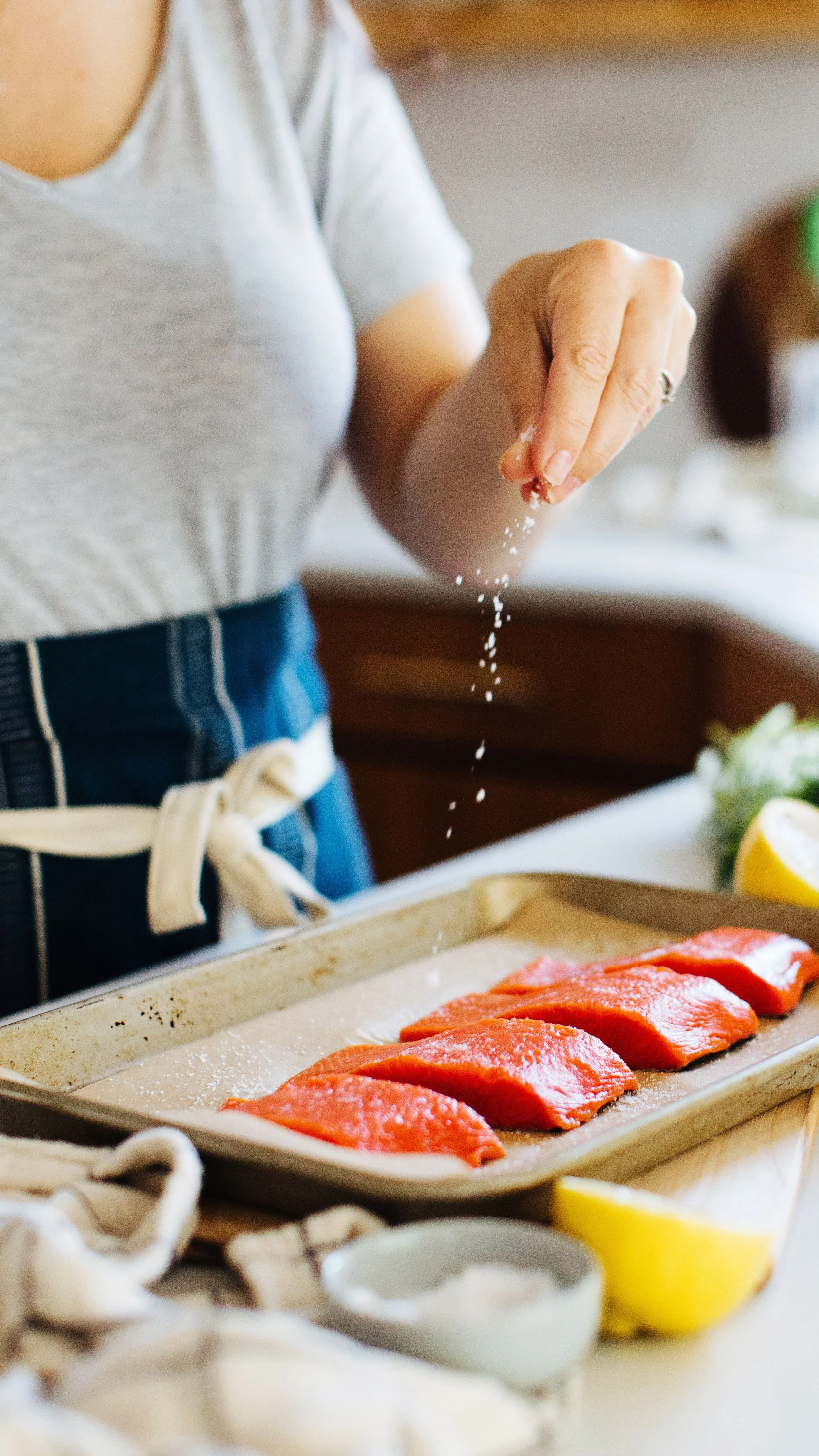 Person sprinkling salt on raw salmon fillets on a baking sheet in a kitchen.