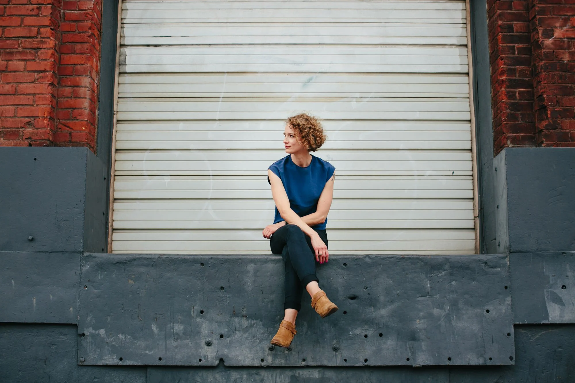 A woman with curly blonde hair sitting on a ledge in front of a closed white roll-up garage door, with red brick walls on either side, wearing a blue sleeveless top and brown ankle boots.
