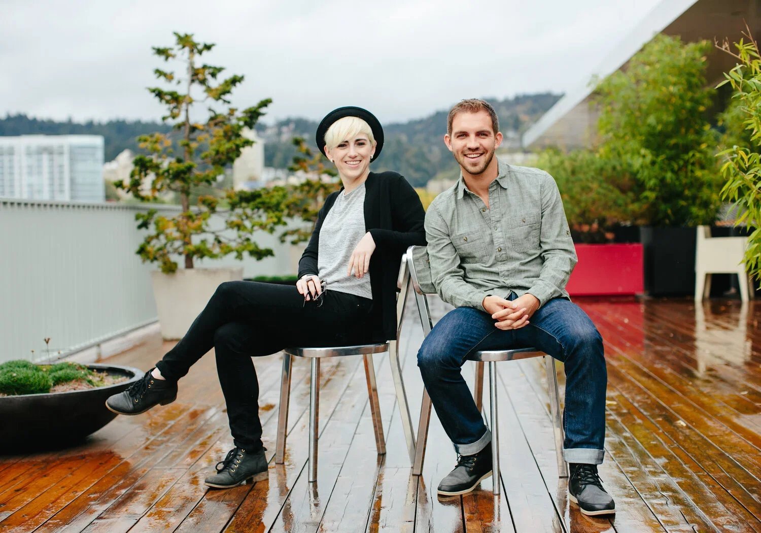 Two young adults sitting and smiling on a rooftop terrace with cityscape and mountains in the background, rainy weather, wooden floor, and potted plants.