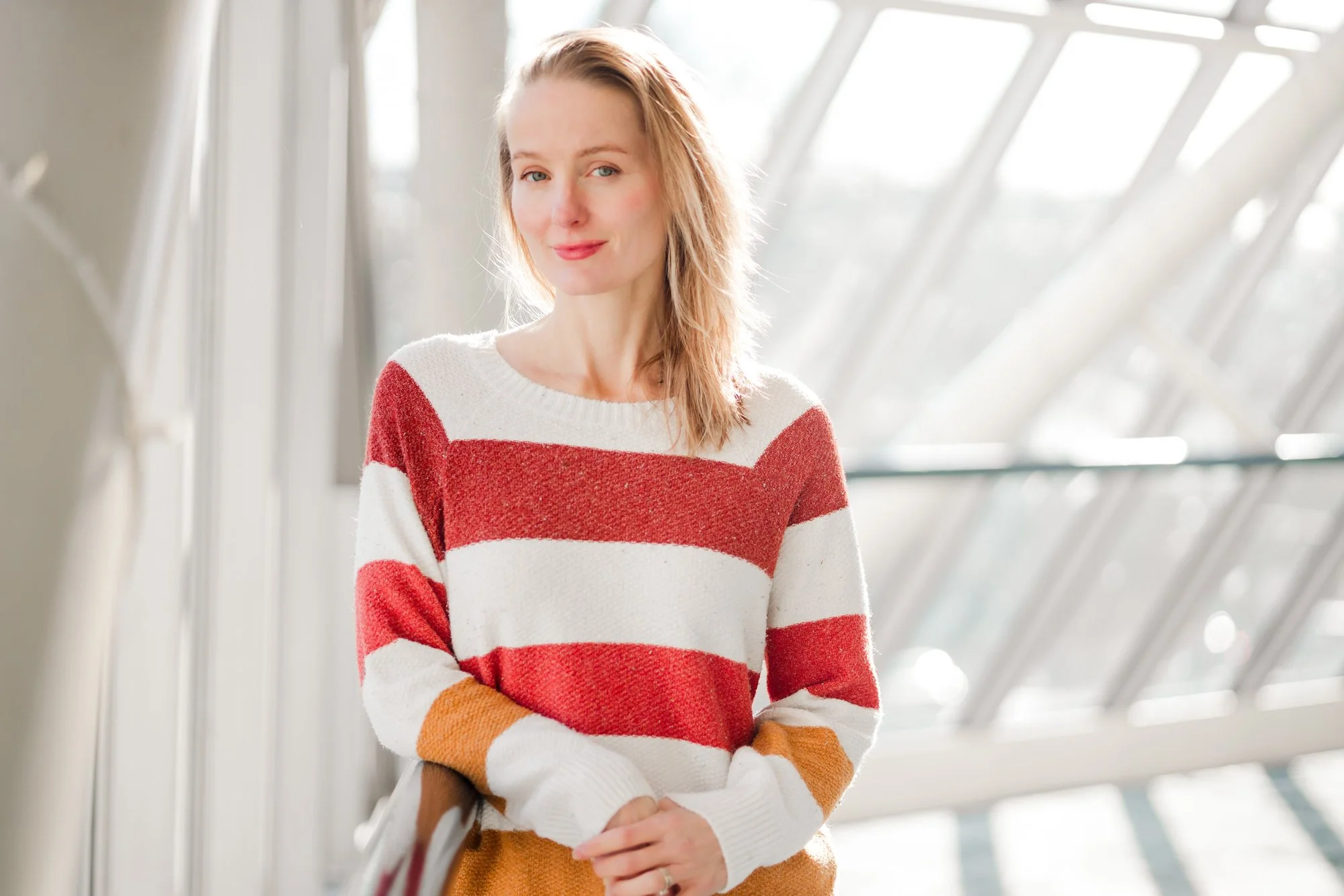 A woman with blonde hair wearing a red and white striped sweater, standing indoors near a window with geometric framing, holding a bag, with sunlight illuminating her face.