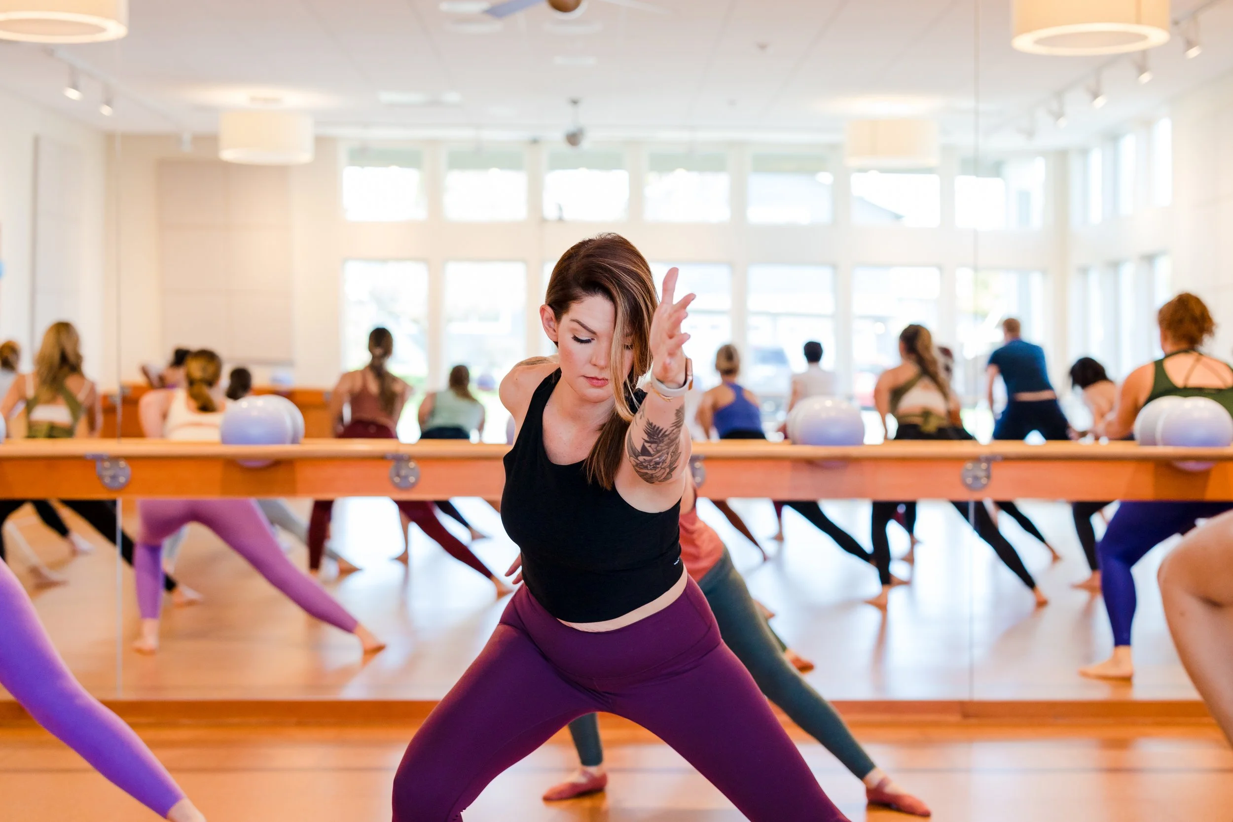 A woman leading a yoga class in a bright studio with large windows, mirror, and other participants doing yoga poses.