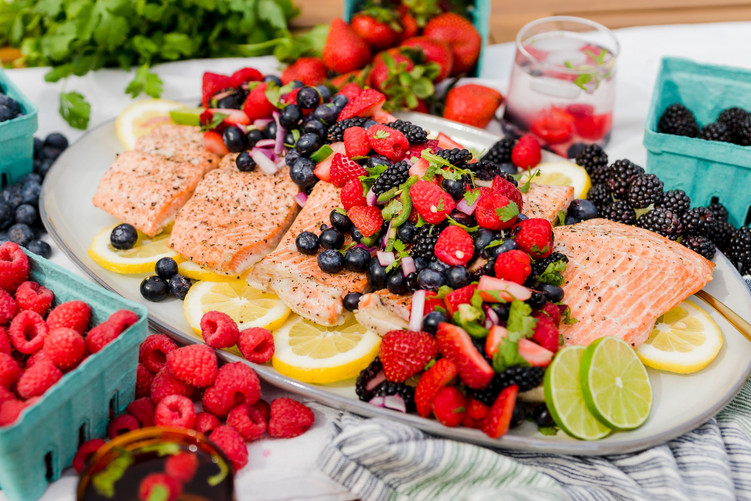 Fresh salmon fillets topped with mixed berries and garnished with lemon slices on a large platter, surrounded by baskets of raspberries, blackberries, and strawberries, with a glass of berry-infused water in the background.