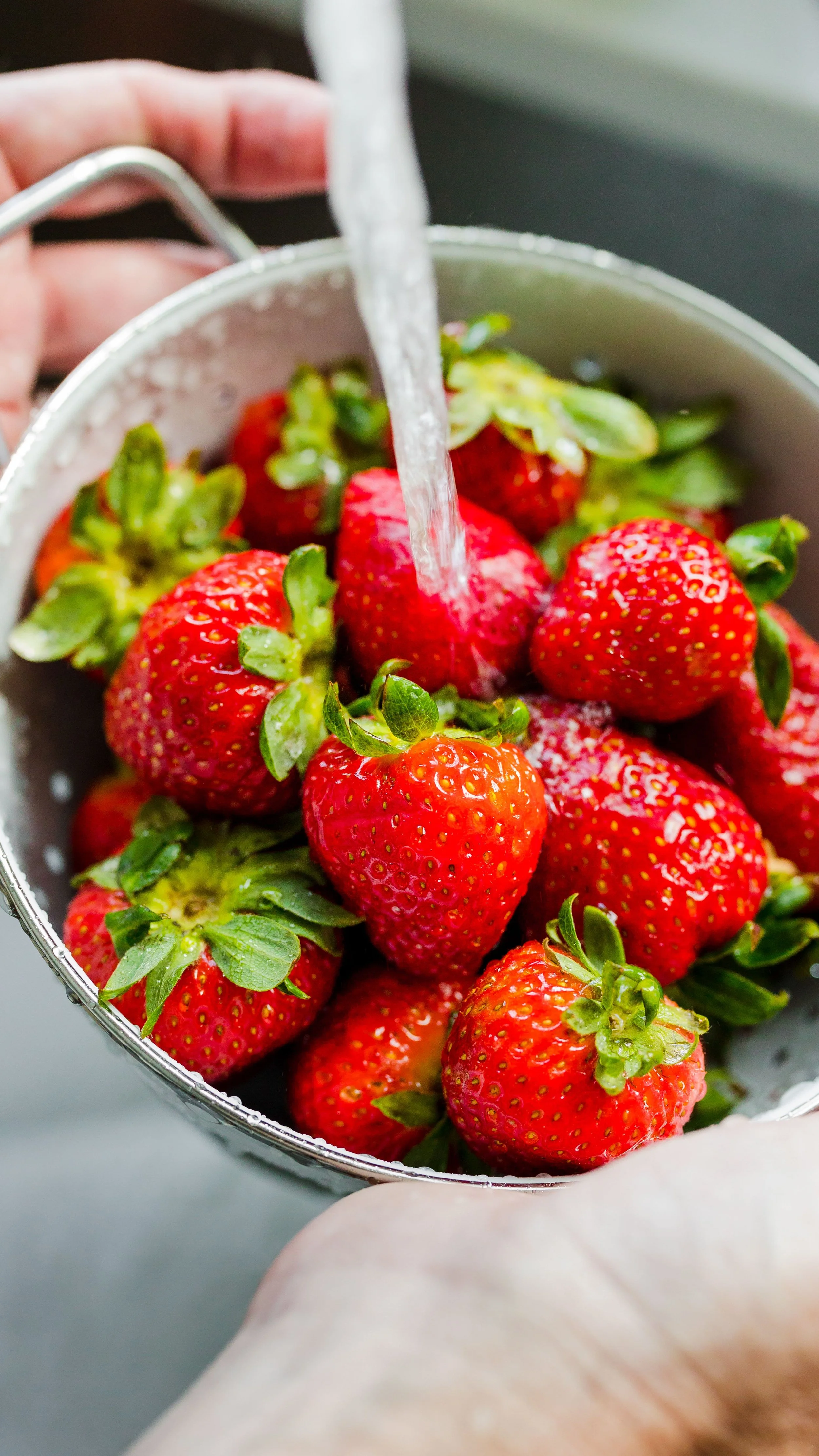 Close-up of fresh strawberries being washed under running water in a metal colander.