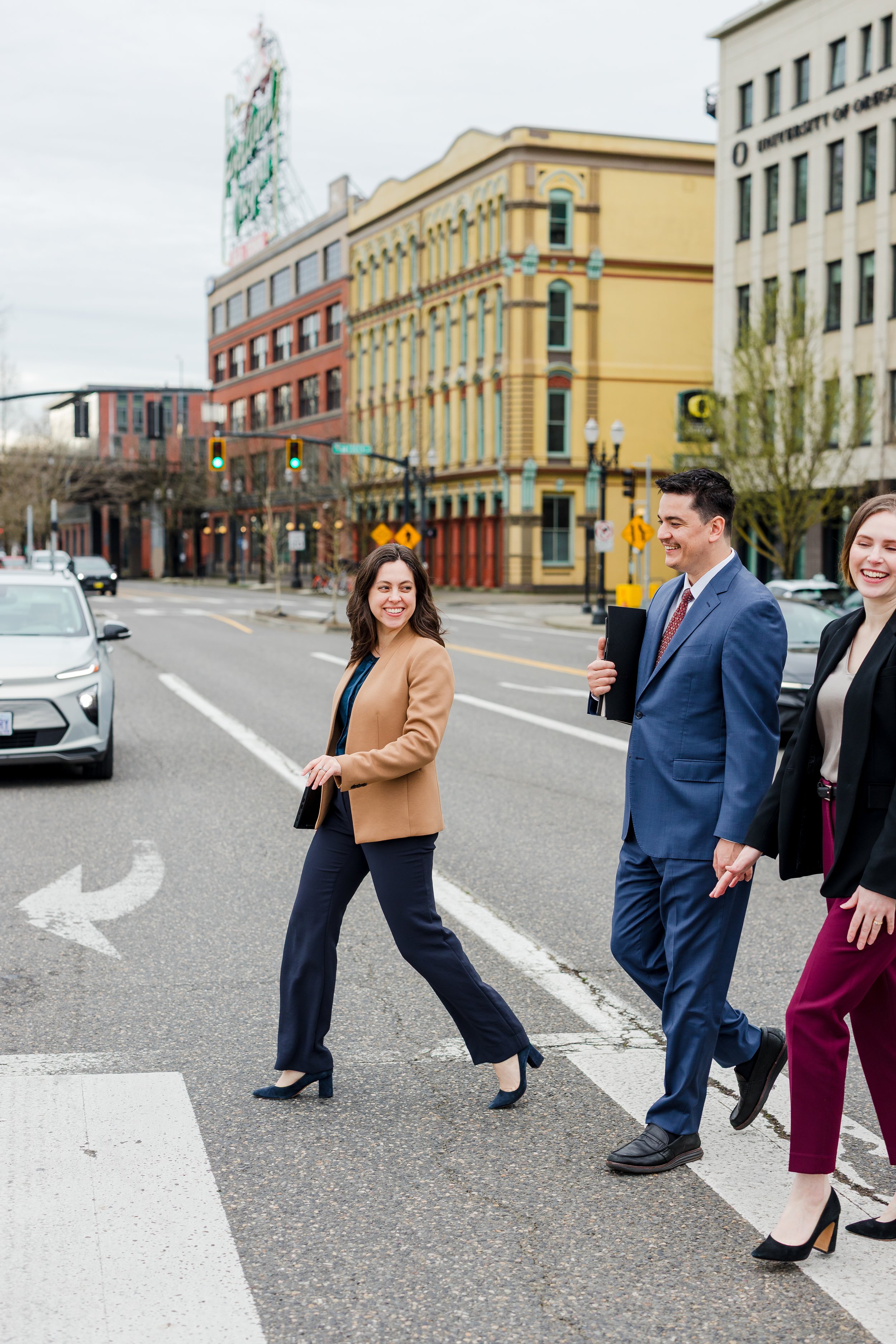 Three business professionals crossing the street in an urban area, smiling and carrying folders, with colorful buildings and traffic lights in the background.