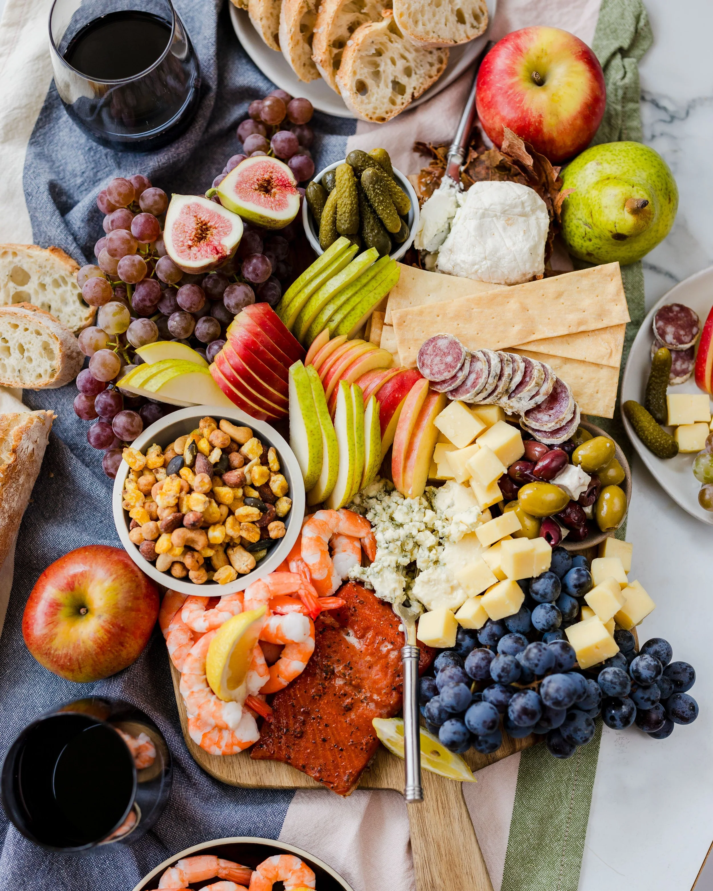A spread of assorted appetizers on a table, including sliced bread, grapes, apples, cheese, cured meats, pickles, shrimp, smoked salmon, and various dips, with glasses of red wine.