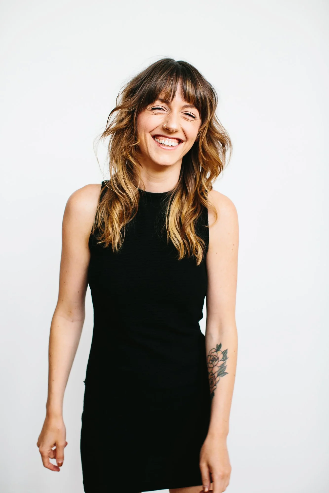 A smiling woman with shoulder-length wavy hair, wearing a black sleeveless dress, standing against a plain white background.