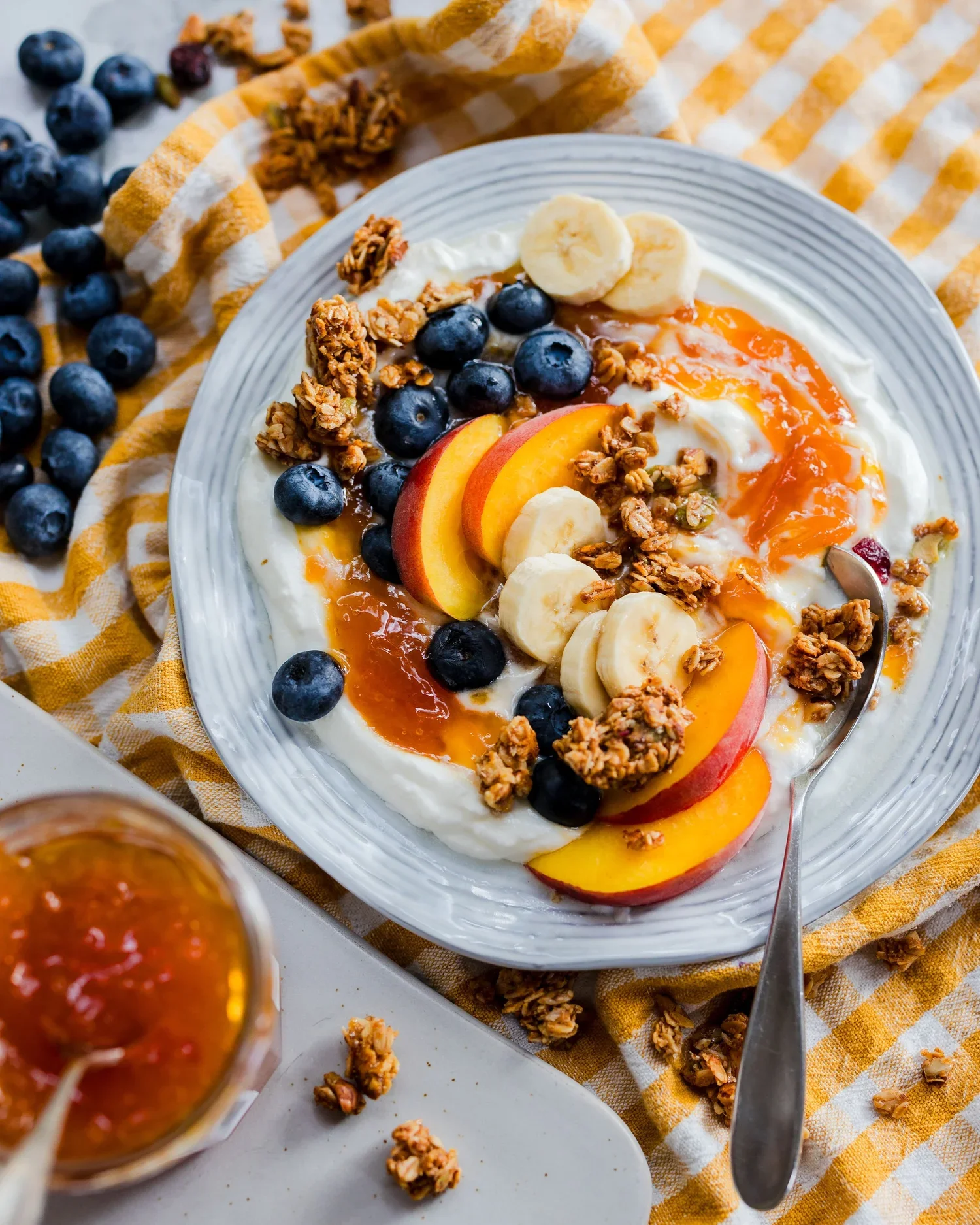 Yogurt parfait topped with blueberries, sliced bananas, peach slices, granola, and drizzled with honey, on a white plate with a fork, surrounded by a yellow and white checkered cloth and a small cup of jam.