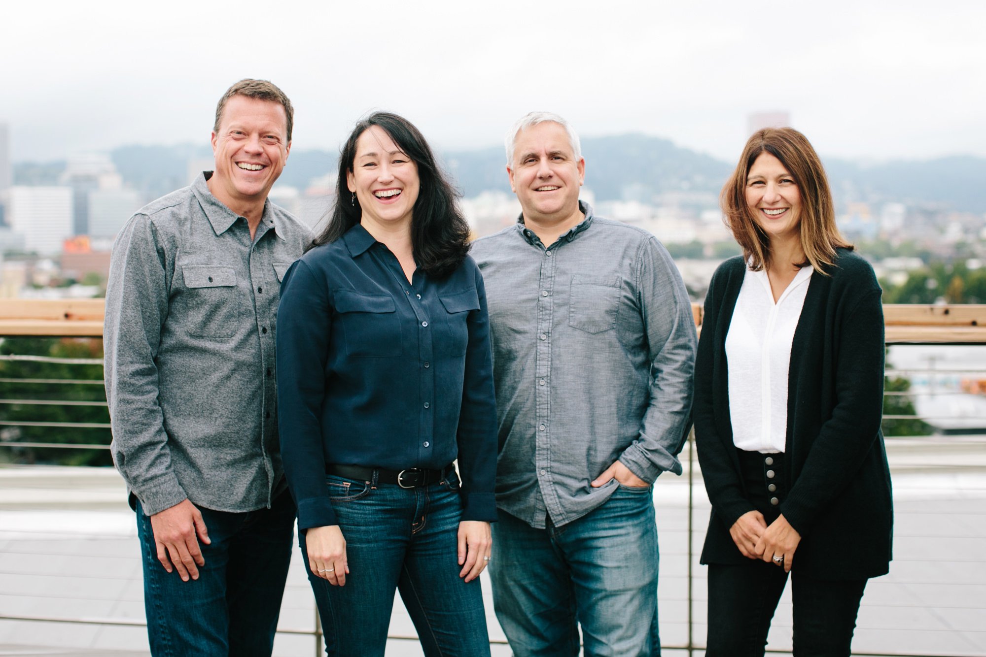 Group of four smiling adults standing outdoors on a balcony with a cityscape in the background.