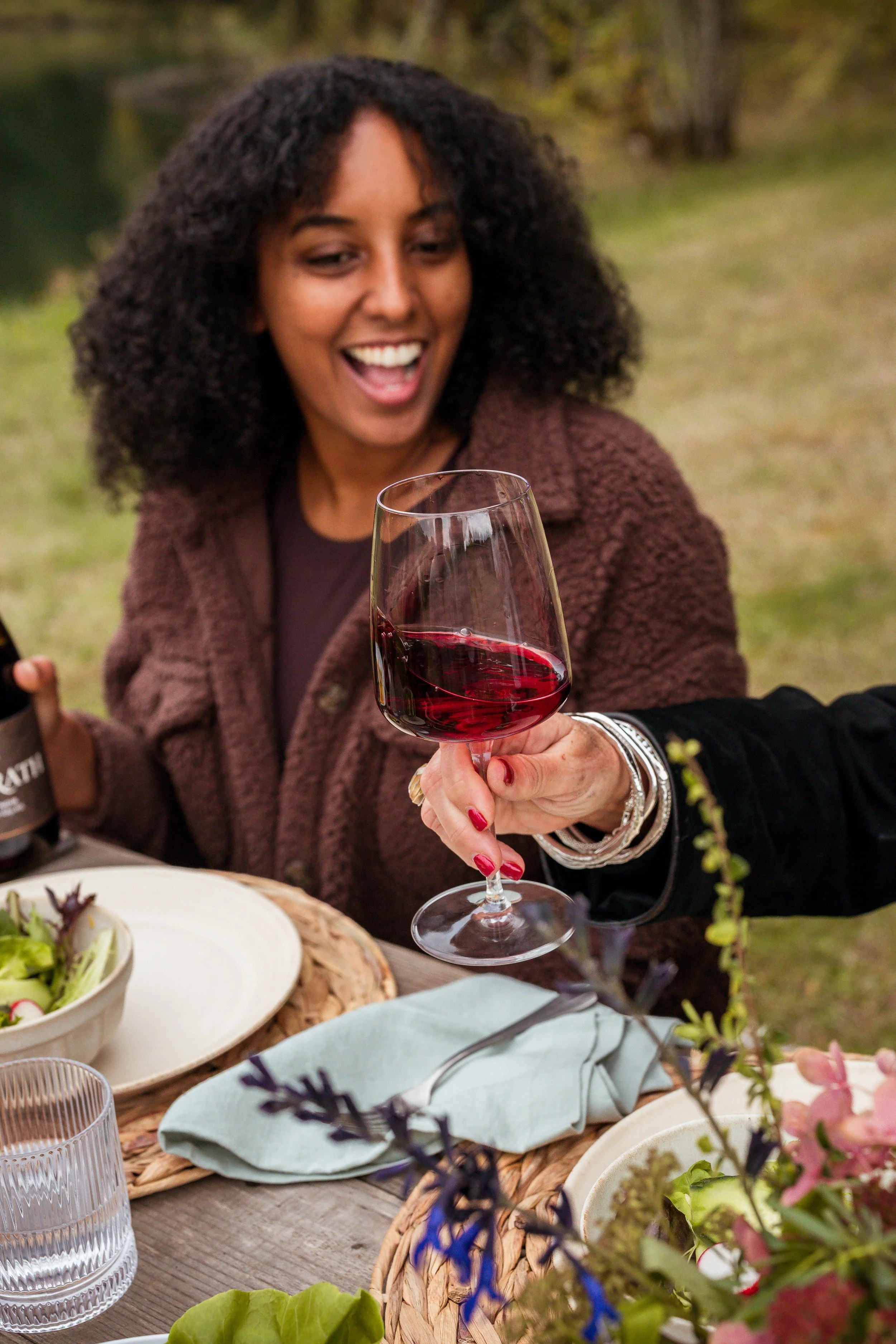 A woman with curly hair, smiling and holding a glass of red wine, sitting at an outdoor table with food and flowers.