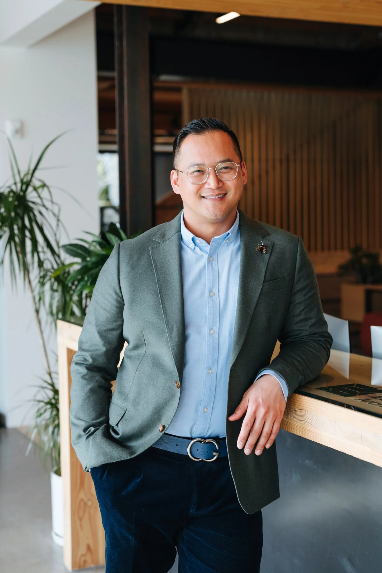 Man in glasses and a gray blazer smiling and leaning on a wooden counter in a modern indoor space with plants and wooden accents.
