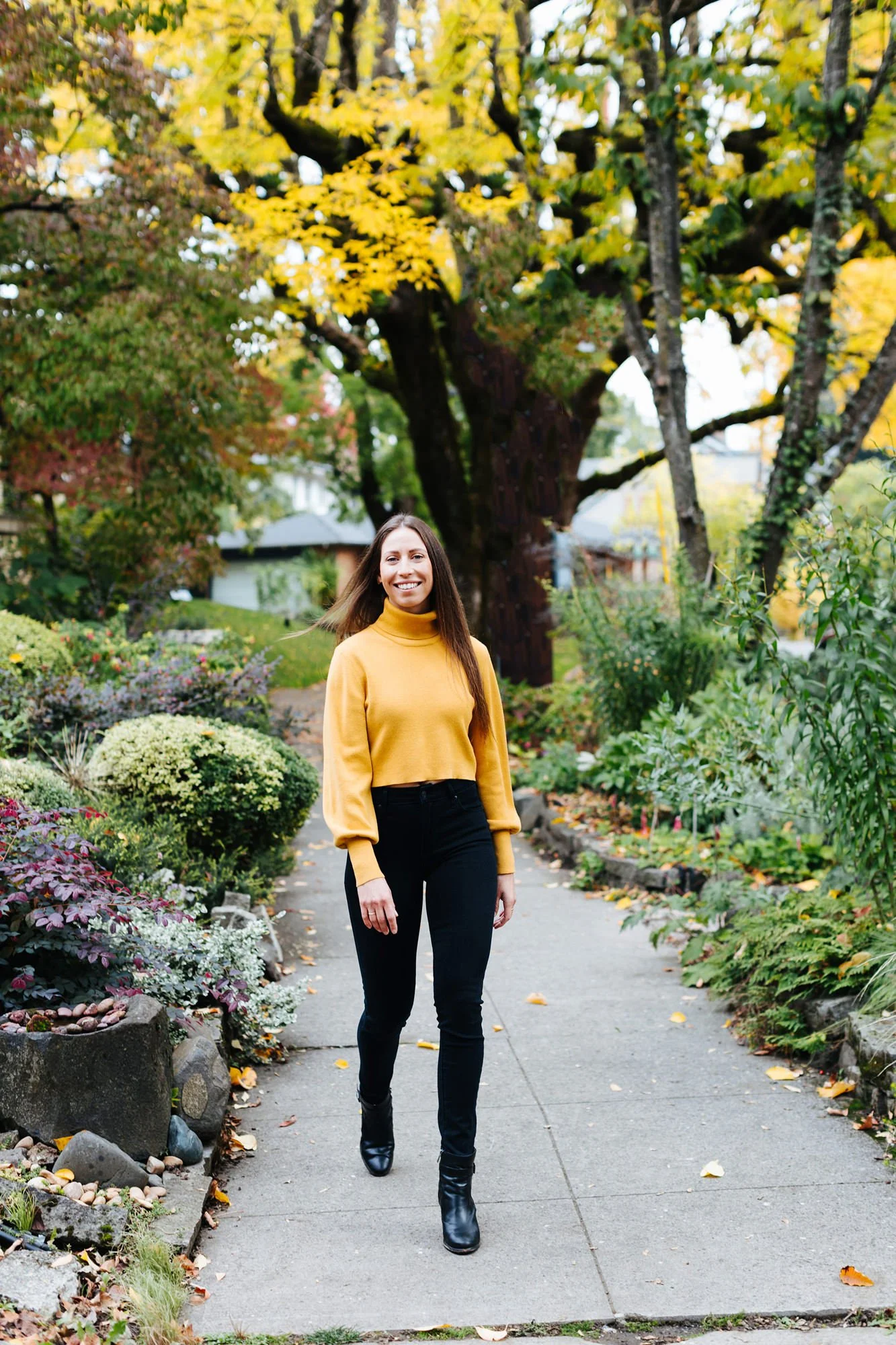 A woman in a yellow sweater and black pants walking on a garden path surrounded by fall foliage.