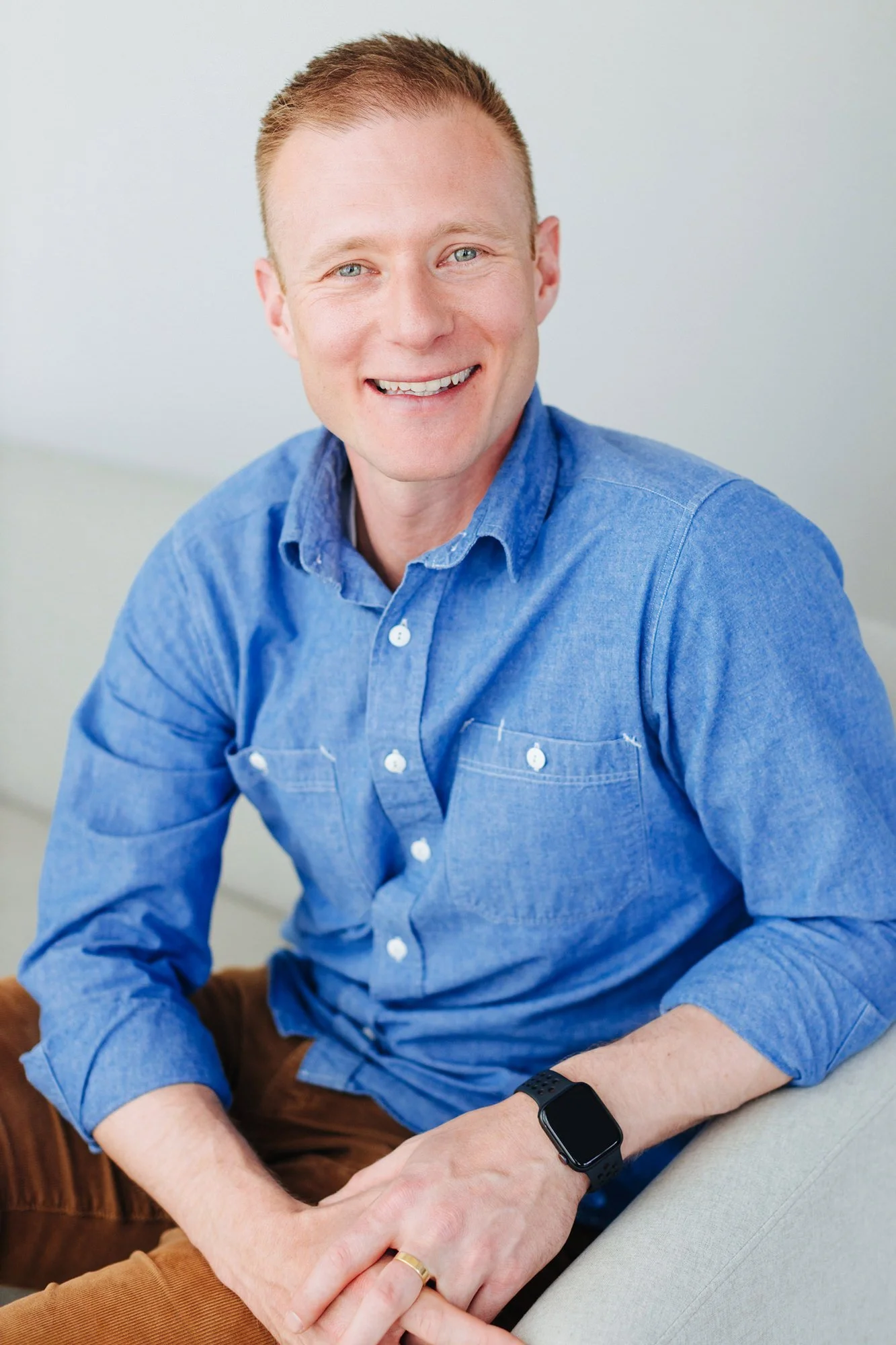 A smiling man with short red hair, light blue eyes, wearing a blue button-up shirt and a black smartwatch, sitting on a light-colored sofa.