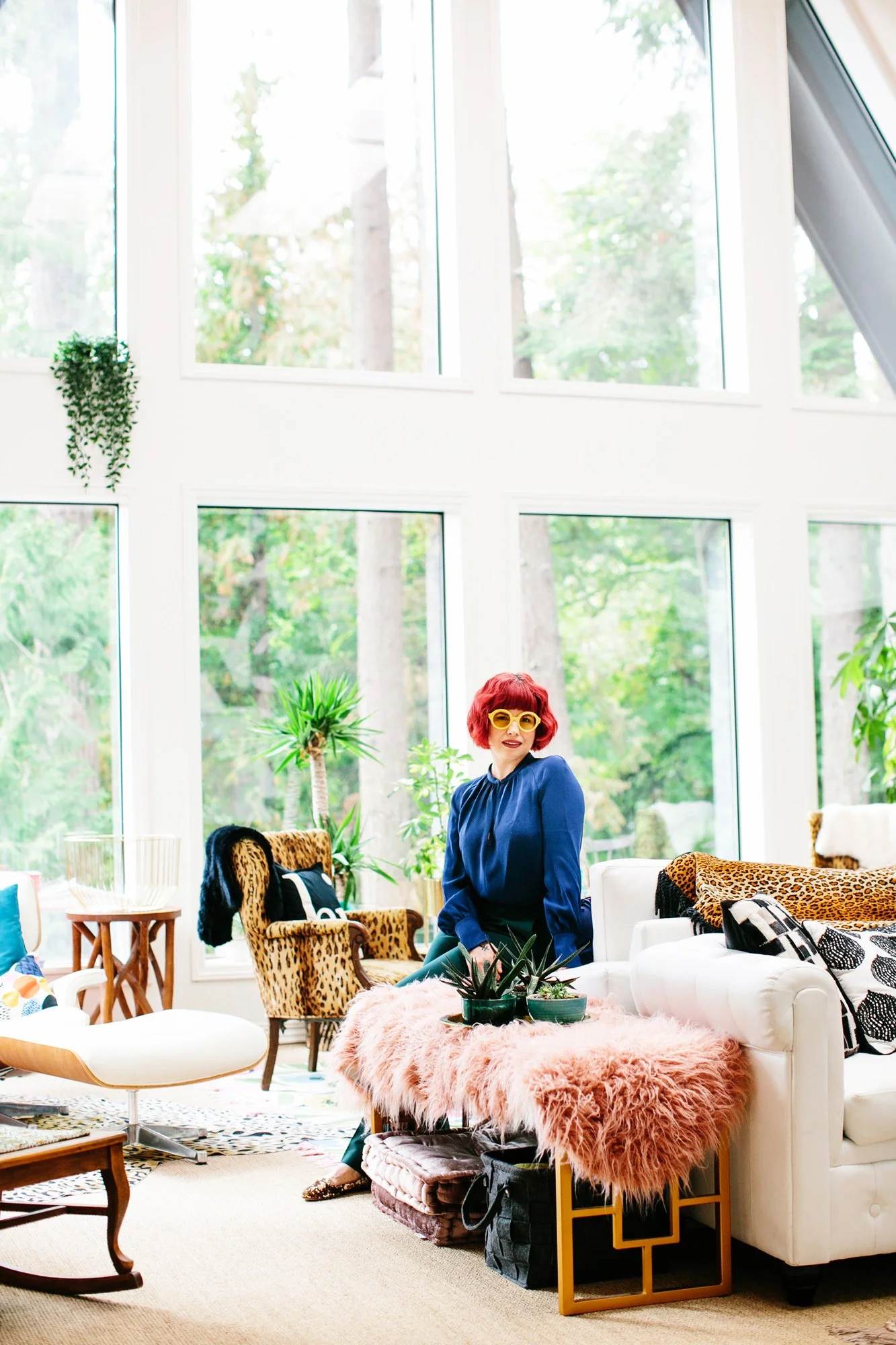 Woman with red hair and yellow glasses sitting by a white sofa in a bright living room with large windows, plants, and eclectic furniture.