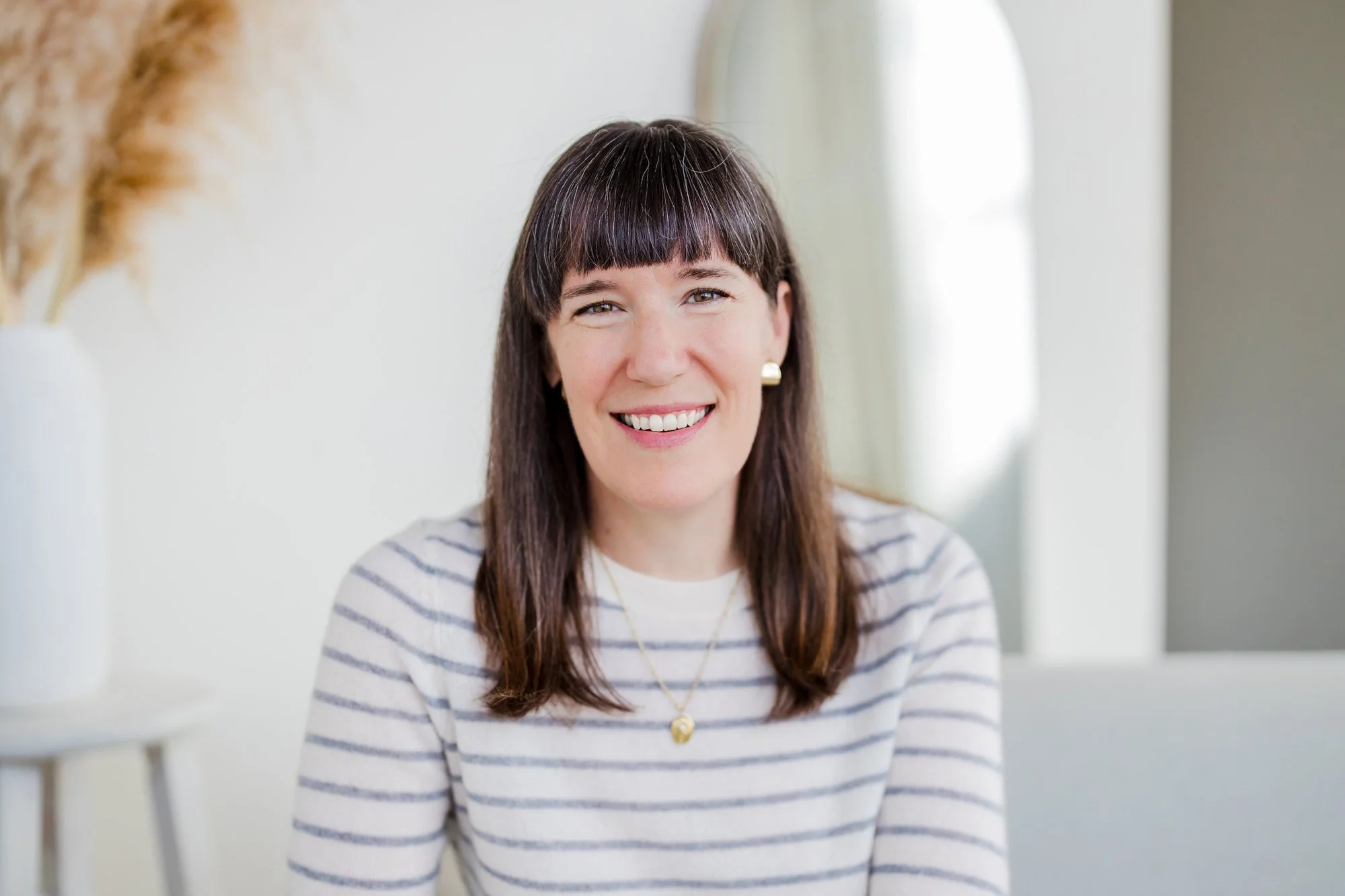A woman with shoulder-length brown hair and bangs, smiling, wearing a white and gray striped shirt, in a bright room with a white background and a large window.