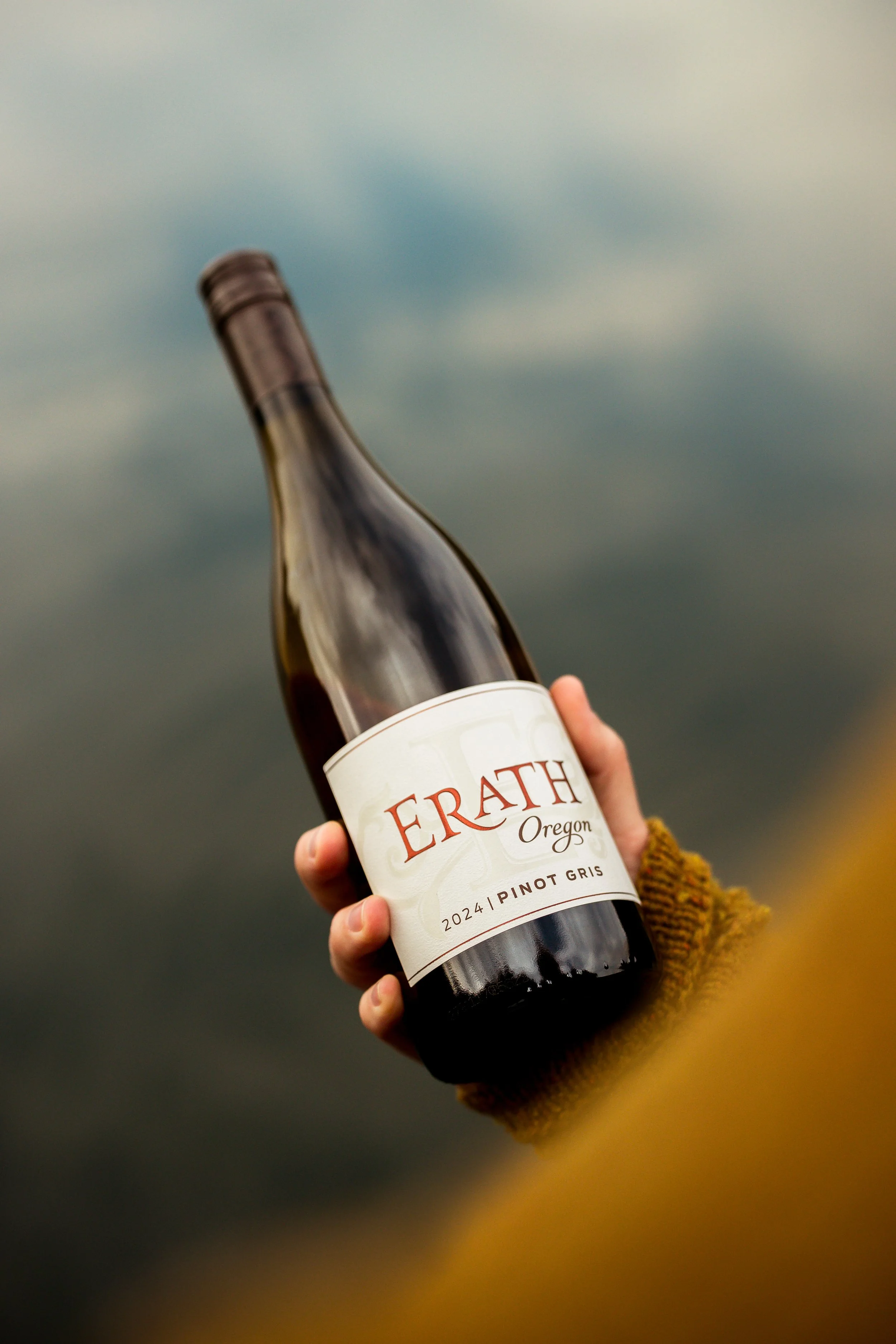 Person holding a bottle of Erath Pinot Gris wine, year 2024, Oregon, with a blurry outdoor background.