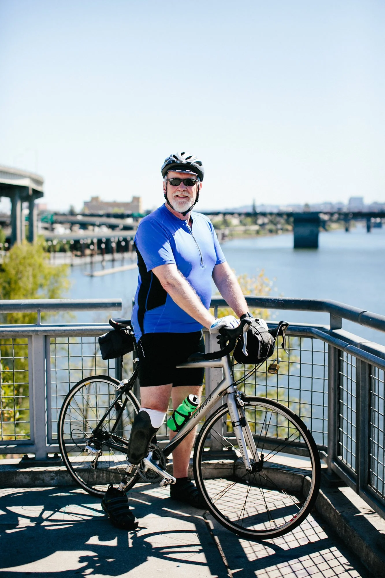 An older man wearing sunglasses and a blue cycling jersey stands with a bicycle on a bridge overlooking a river, with a bridge and cityscape in the background.
