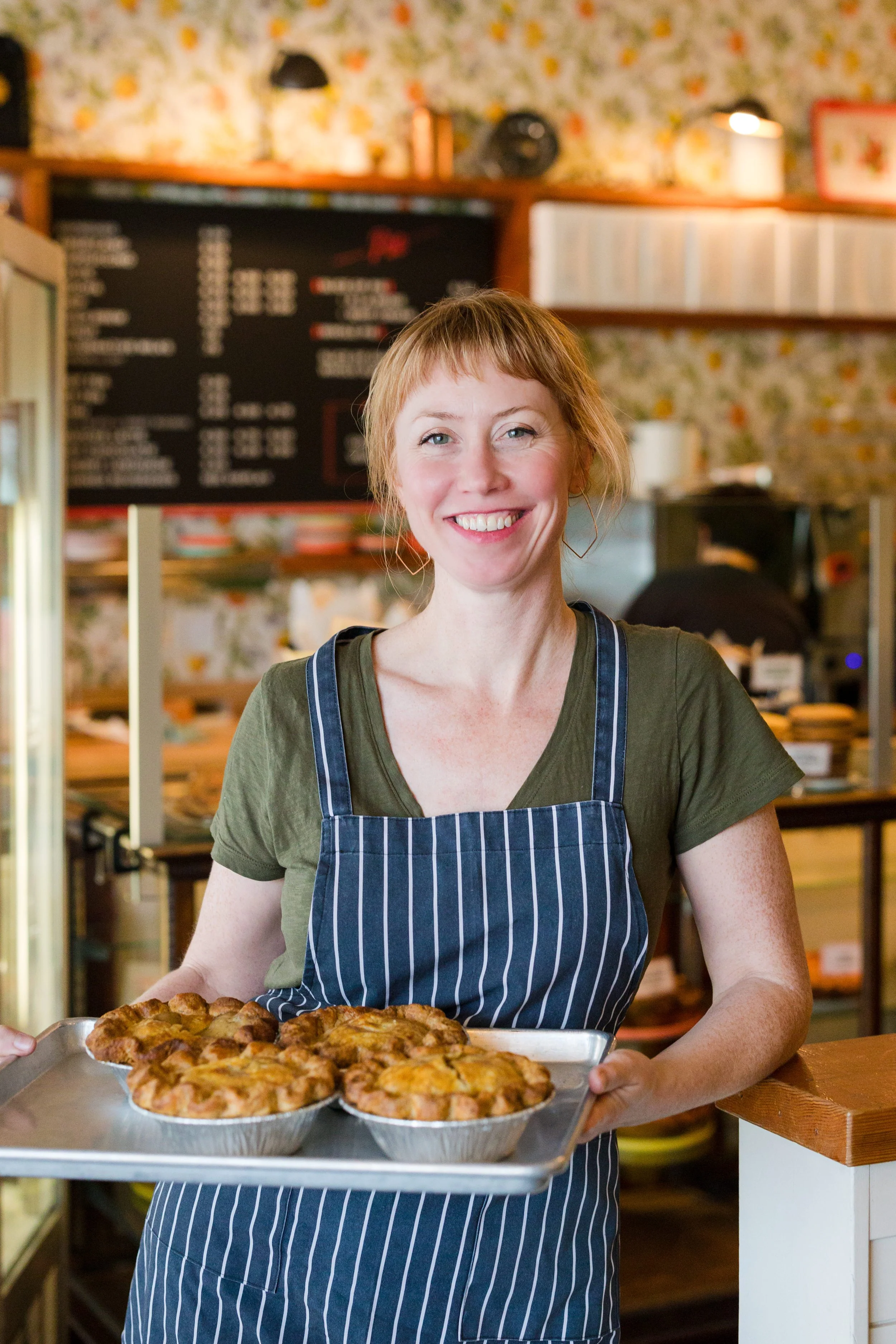 A woman in a green shirt and blue striped apron smiling while holding a tray of freshly baked apple pies inside a bakery.