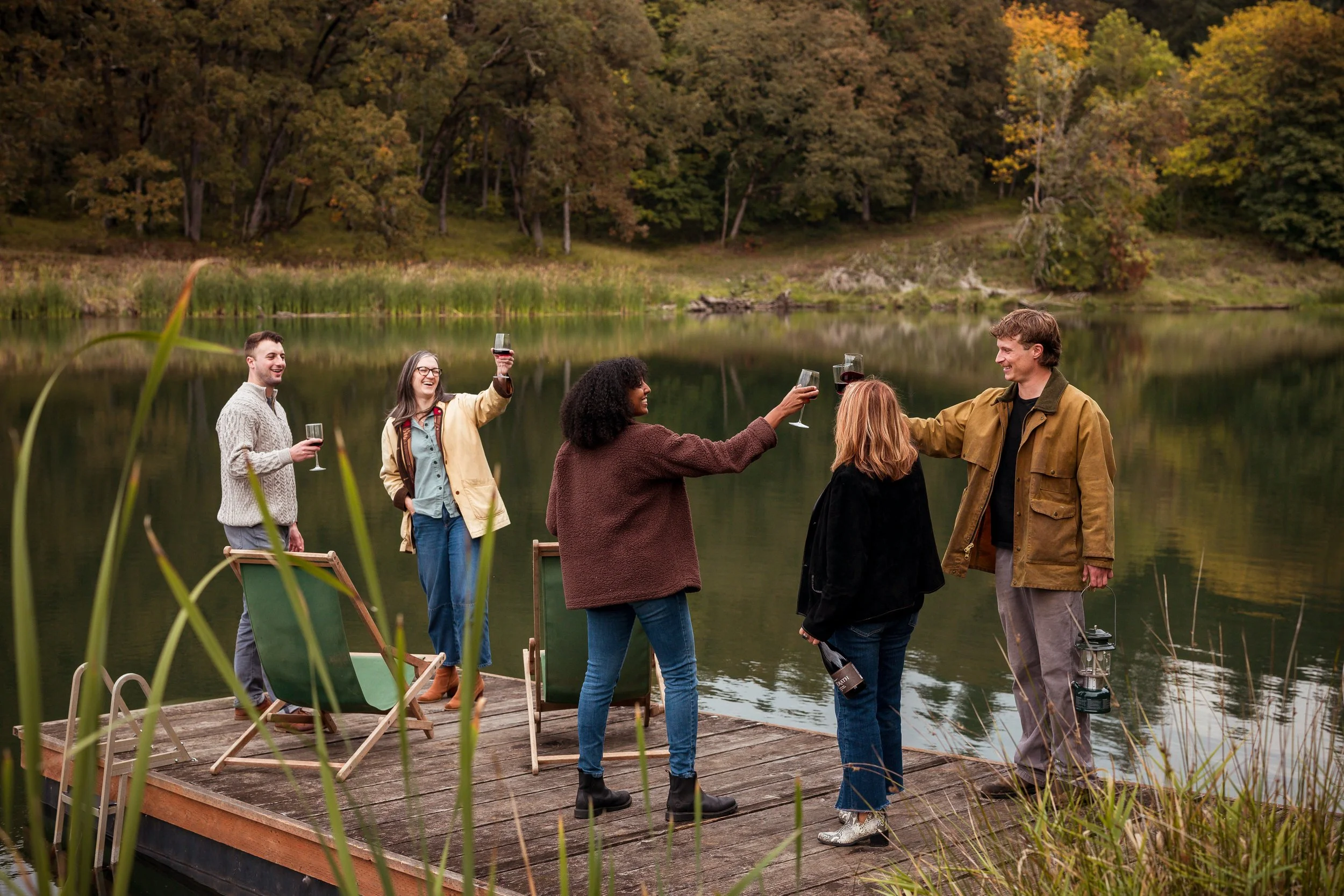 A group of six people standing on a wooden dock by a lake, raising glasses of red wine in a toast, surrounded by trees with autumn foliage.