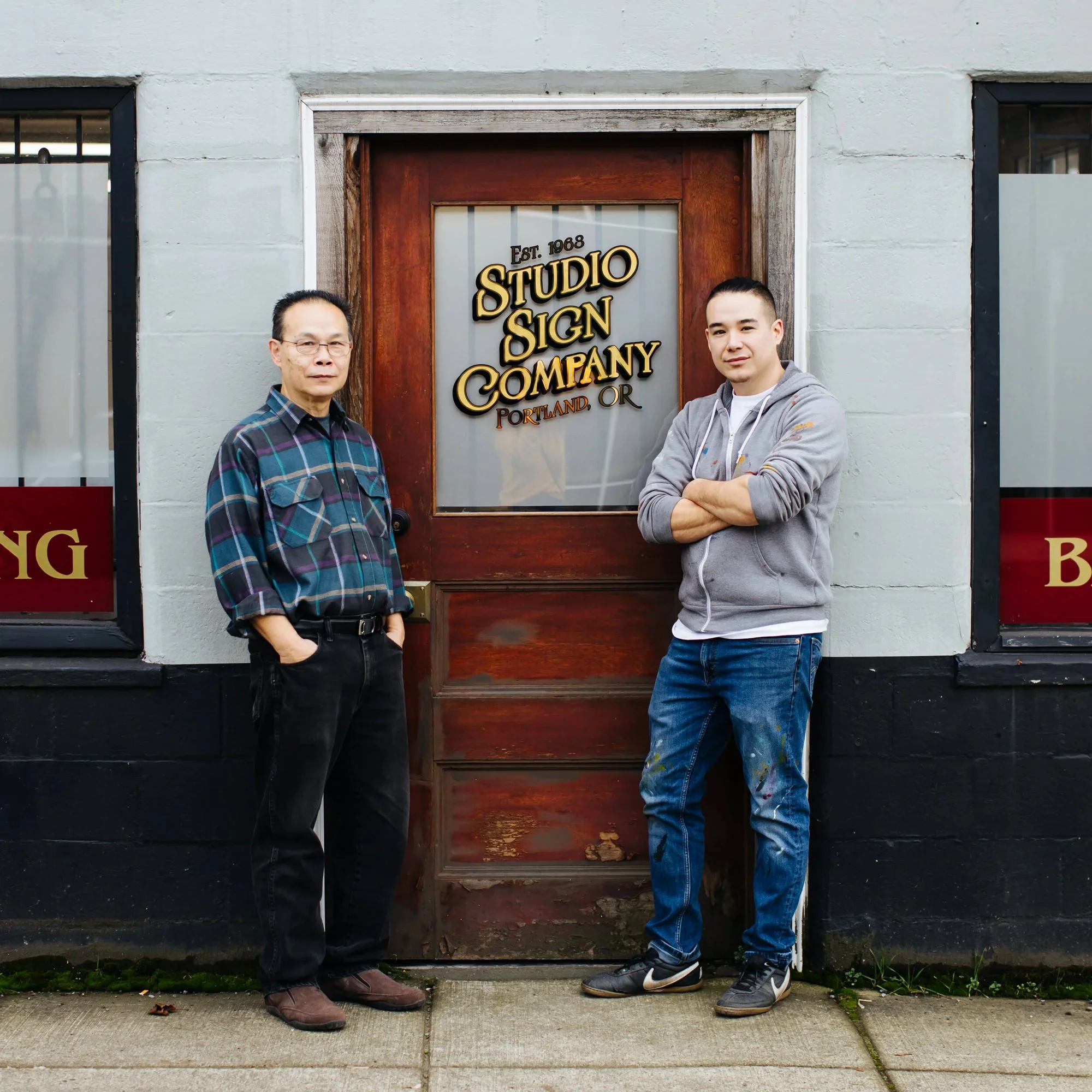 Two men standing in front of a wooden door with a sign that reads "Studio Sign Company, Portland, OR," in front of a gray building.