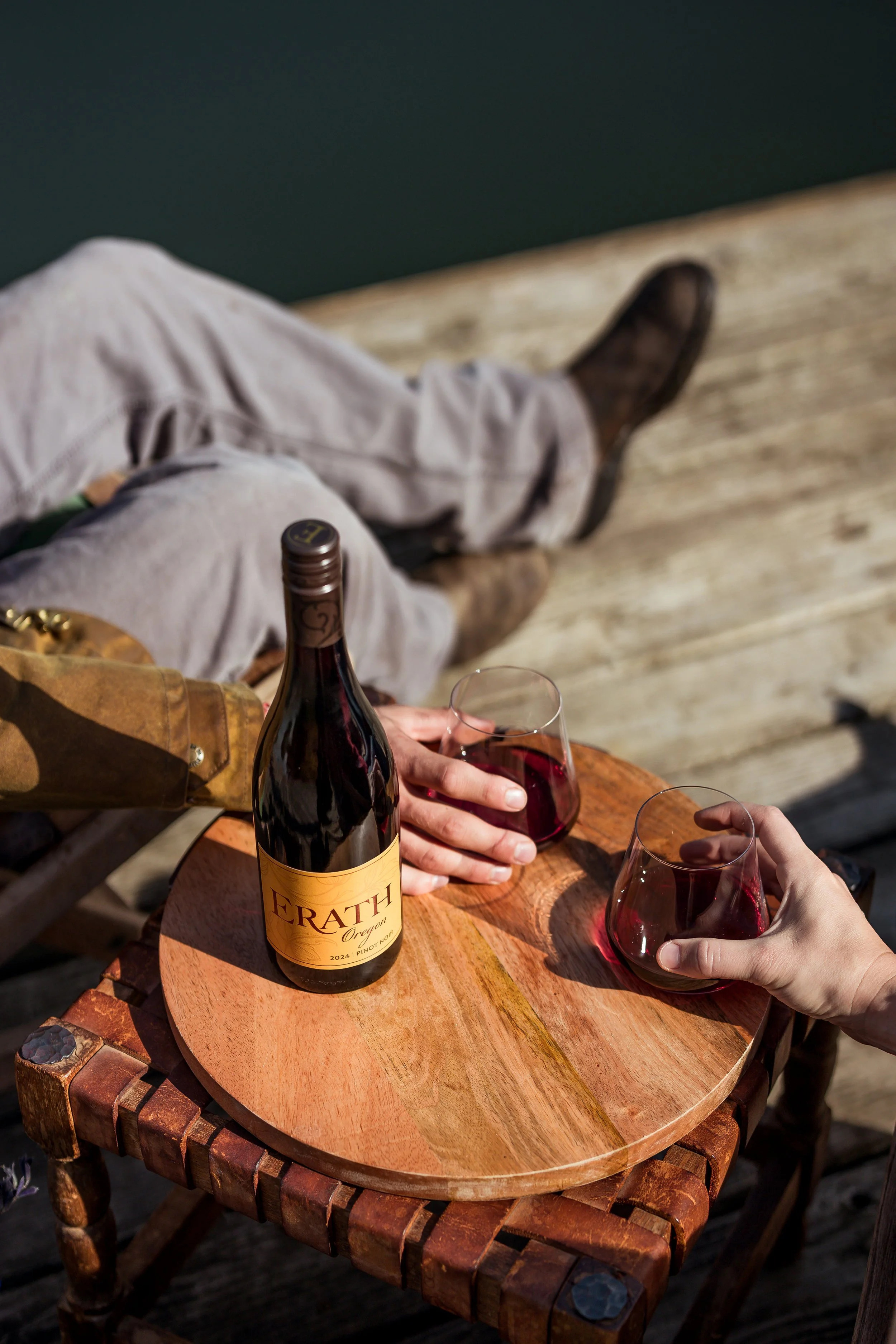Two people holding glasses of red wine over a small wooden table near a body of water. A bottle of Erath Oregon Pinot Noir is on the table, and one person is sitting on a wooden dock.