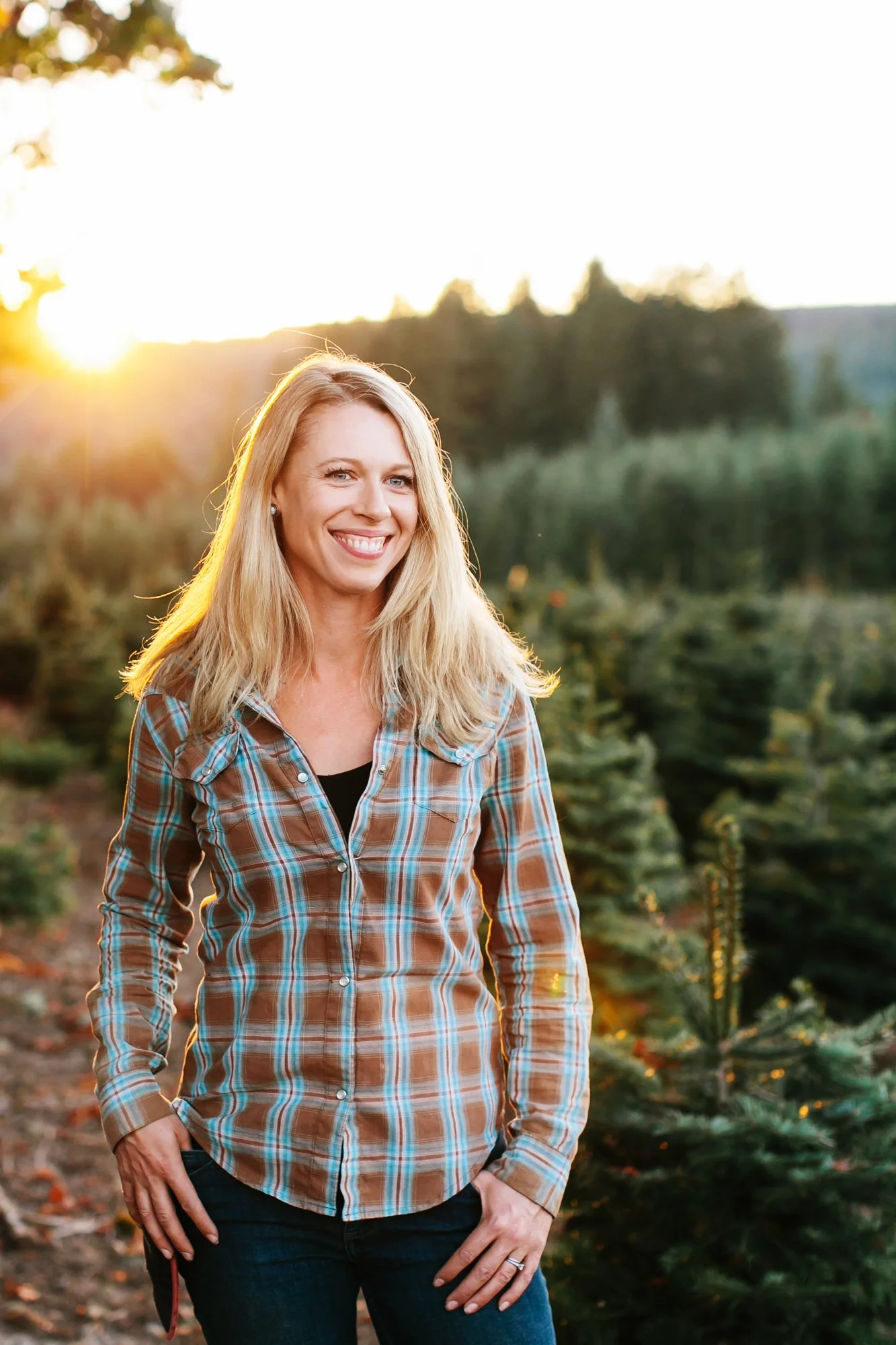 A woman with blonde hair smiling, wearing a plaid shirt, standing outdoors at a Christmas tree farm during sunset.