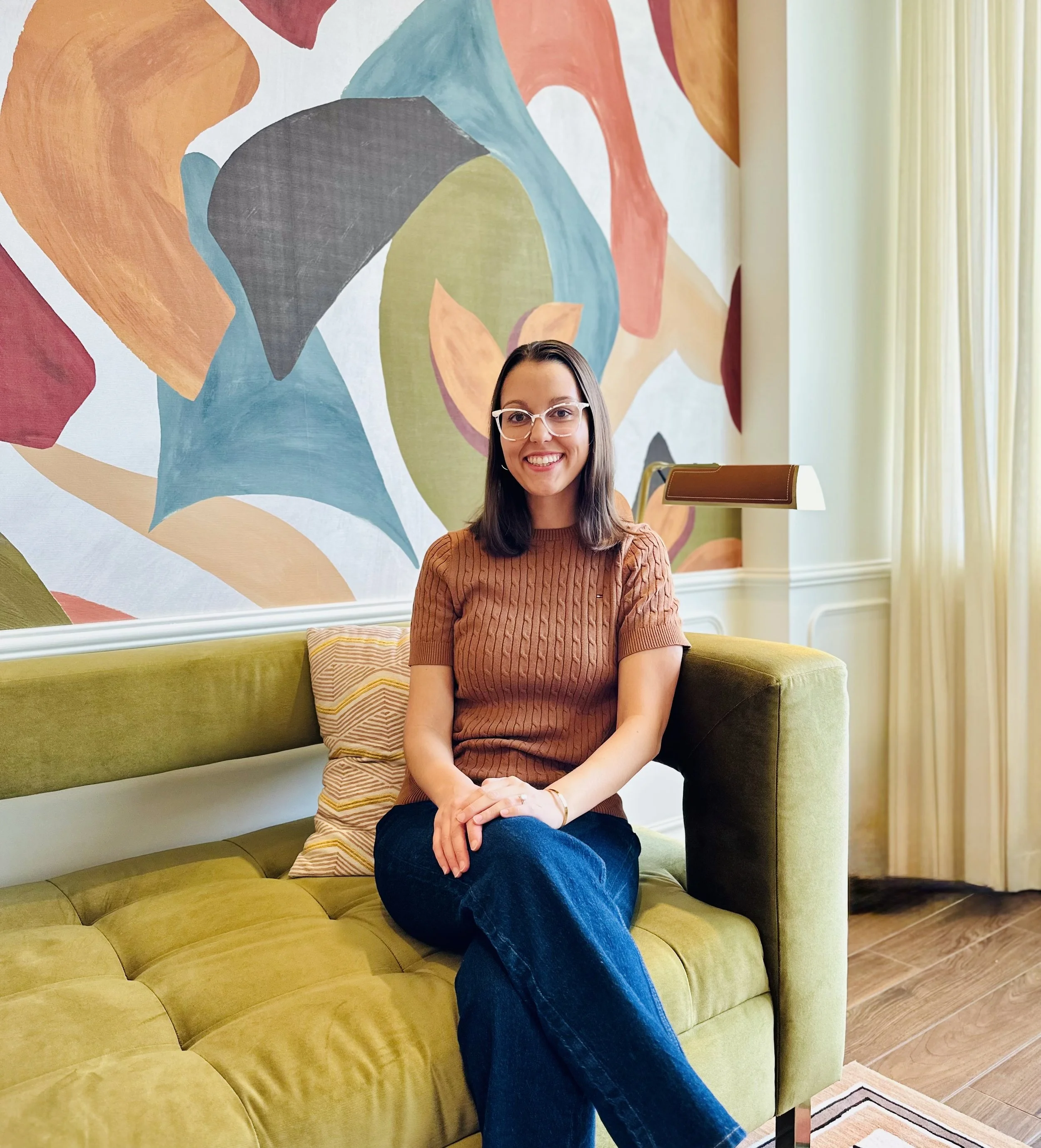 Dr. Hadorn sitting on a couch with legs crossed and hands resting on legs. Abstract mural painted on the wall behind her.