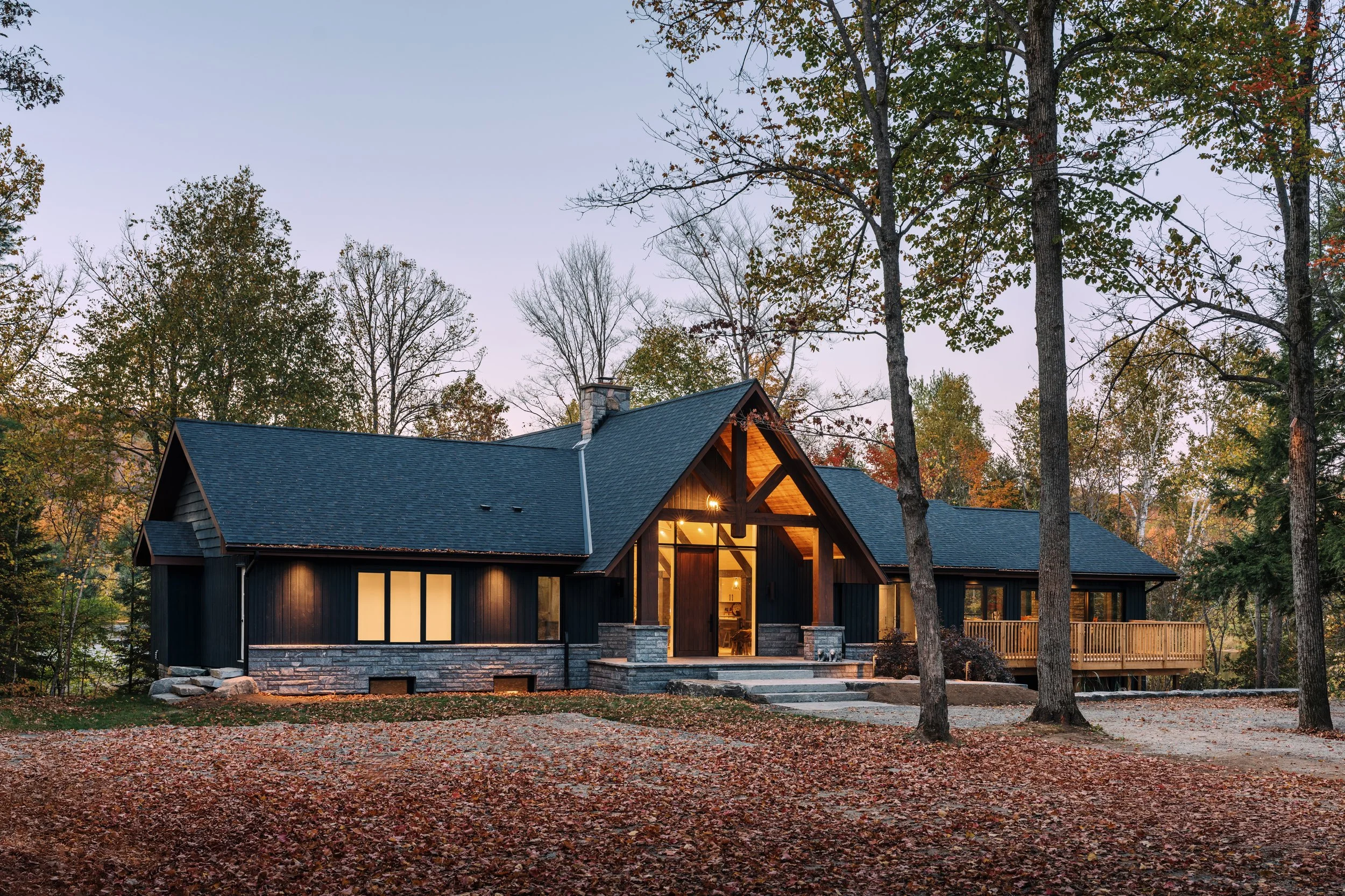 A modern house with black exterior, large windows, and a peaked roof surrounded by trees with fall foliage, during dusk.