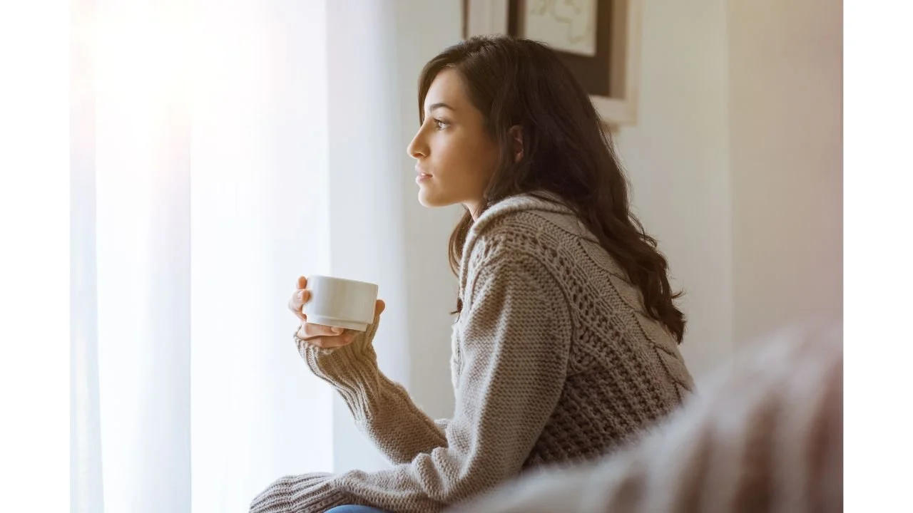 Single mother sitting by a window holding a mug, looking thoughtful and calm in soft morning light.