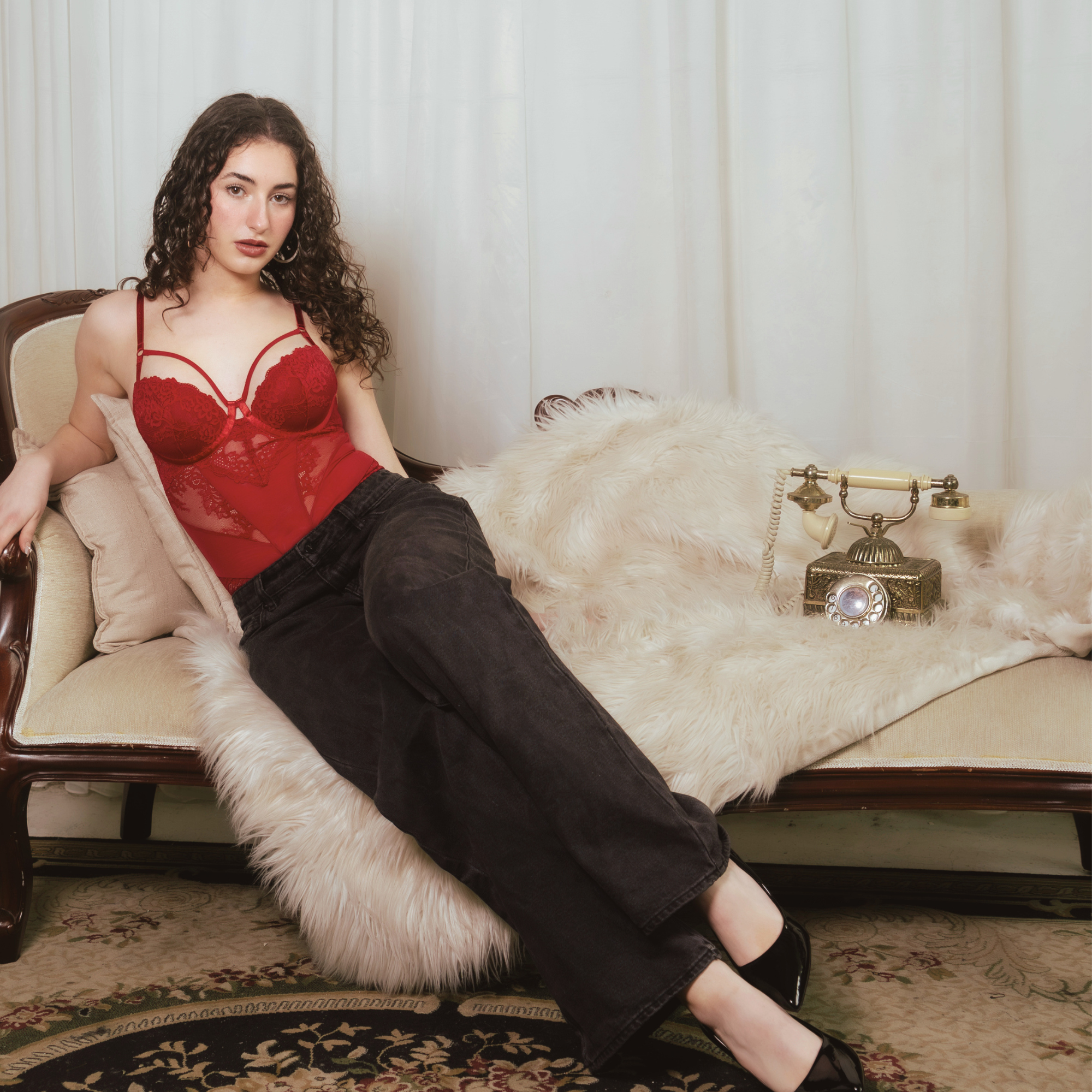 A woman with curly hair wearing a red lace lingerie top and black pants, sitting on a beige sofa with pillows, next to a fur rug and vintage telephone, in a room with white curtains.