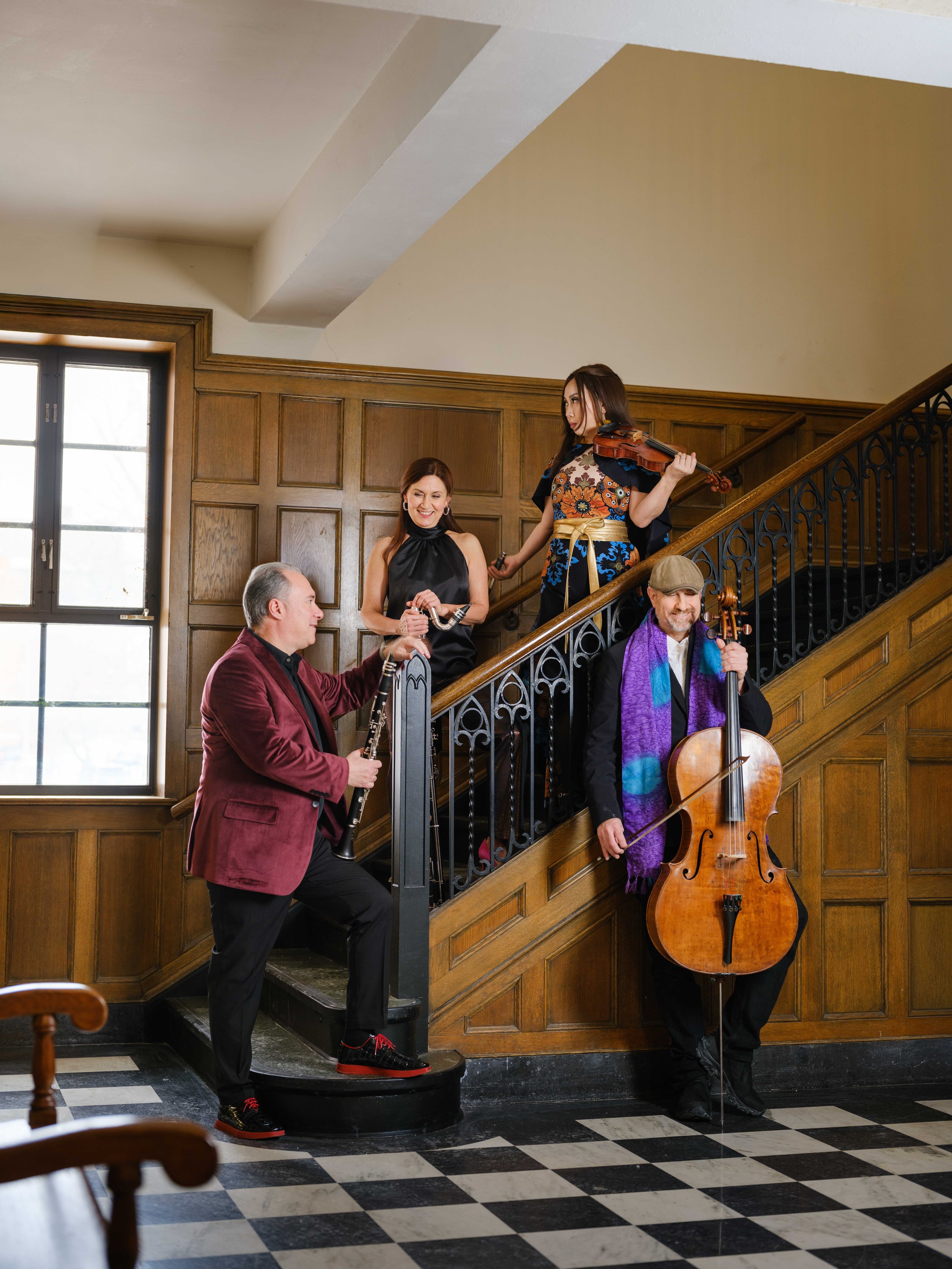 Four musicians holding musical instruments on a wooden staircase in a room with wooden paneled walls and a window.