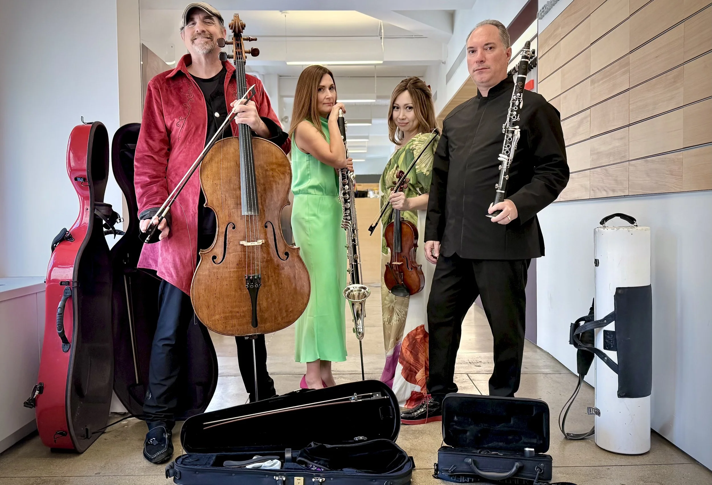 Four musicians posing with their instruments, including a cellist, violinist, flutist, and bassoonist, in a modern indoor setting with instrument cases on the floor.