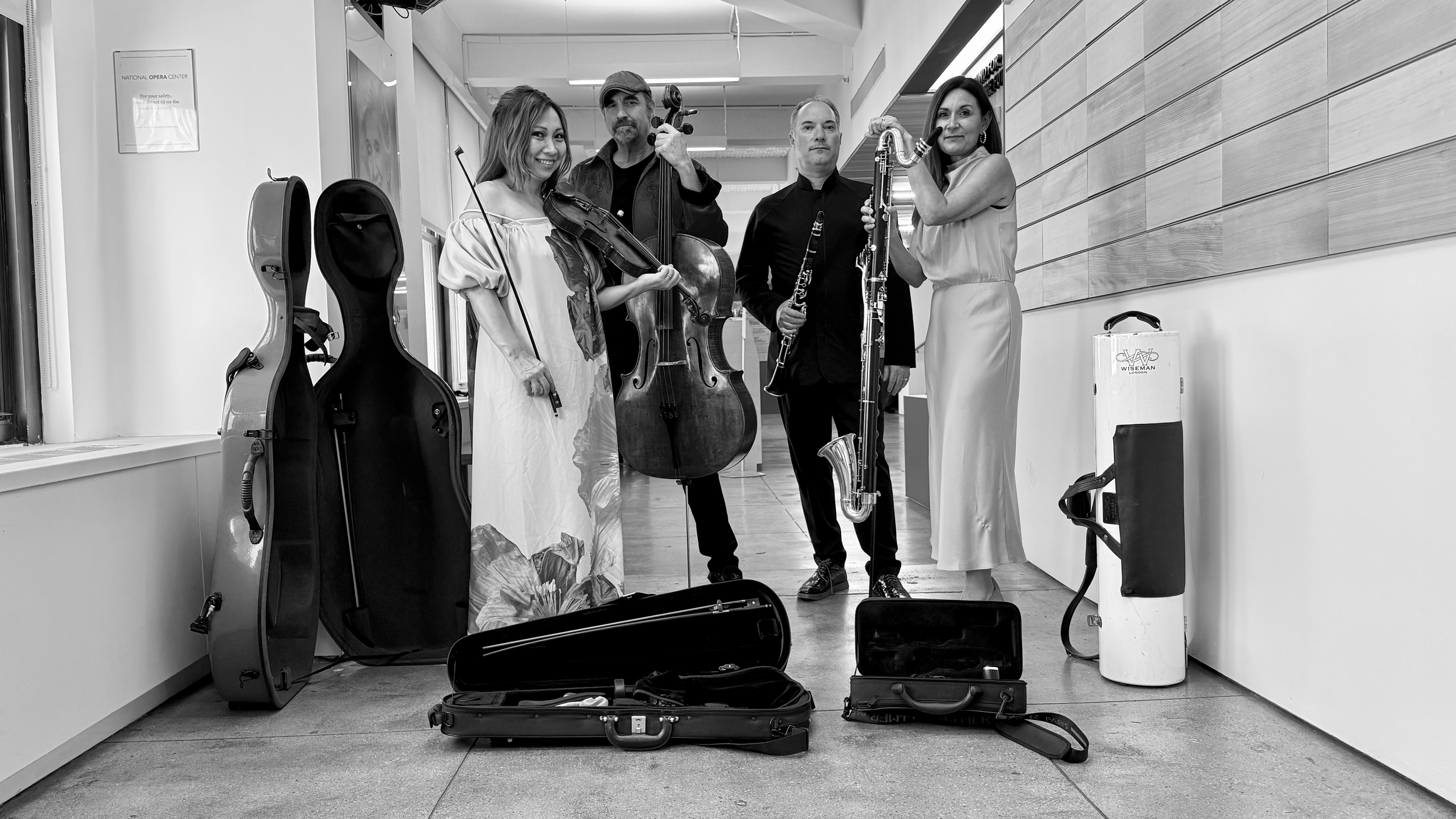 Four musicians holding their instruments in a hallway, with open instrument cases on the floor, and empty instrument cases on the wall behind them.