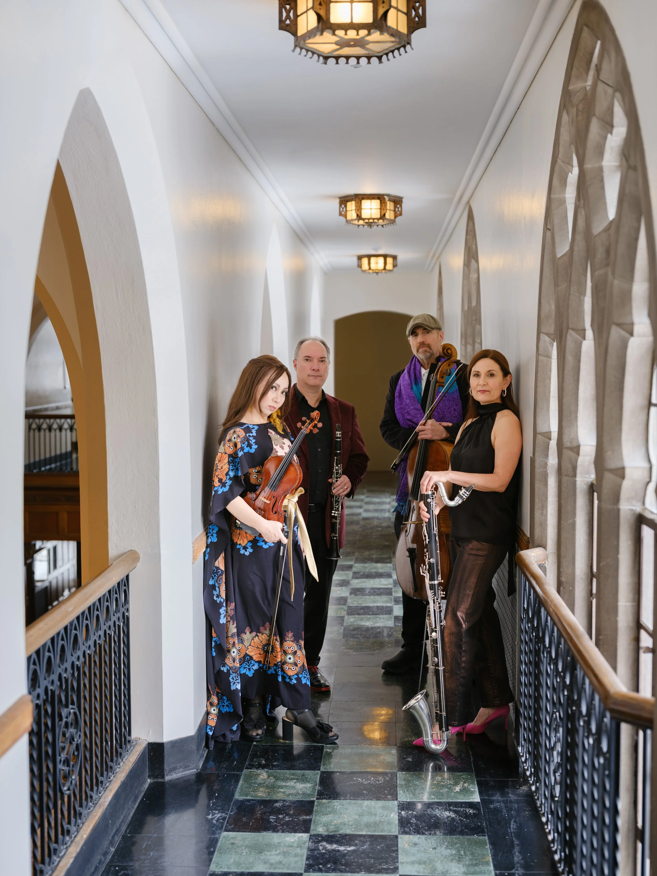 Four musicians with instruments standing in a hallway with arched windows and patterned tile floor.