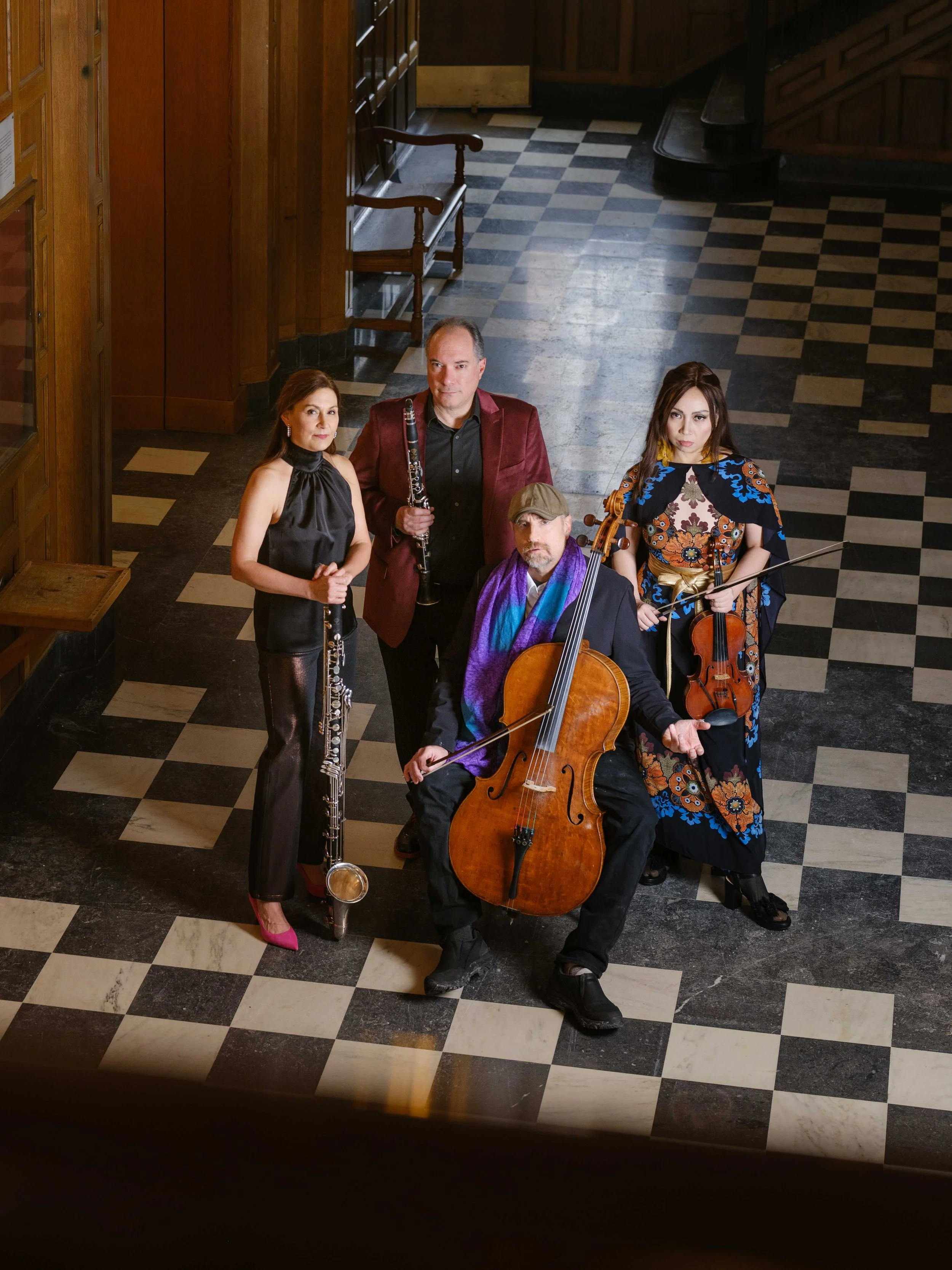 A group of five musicians with string and wind instruments standing inside a wooden-paneled building with a checkered floor, viewed from above.