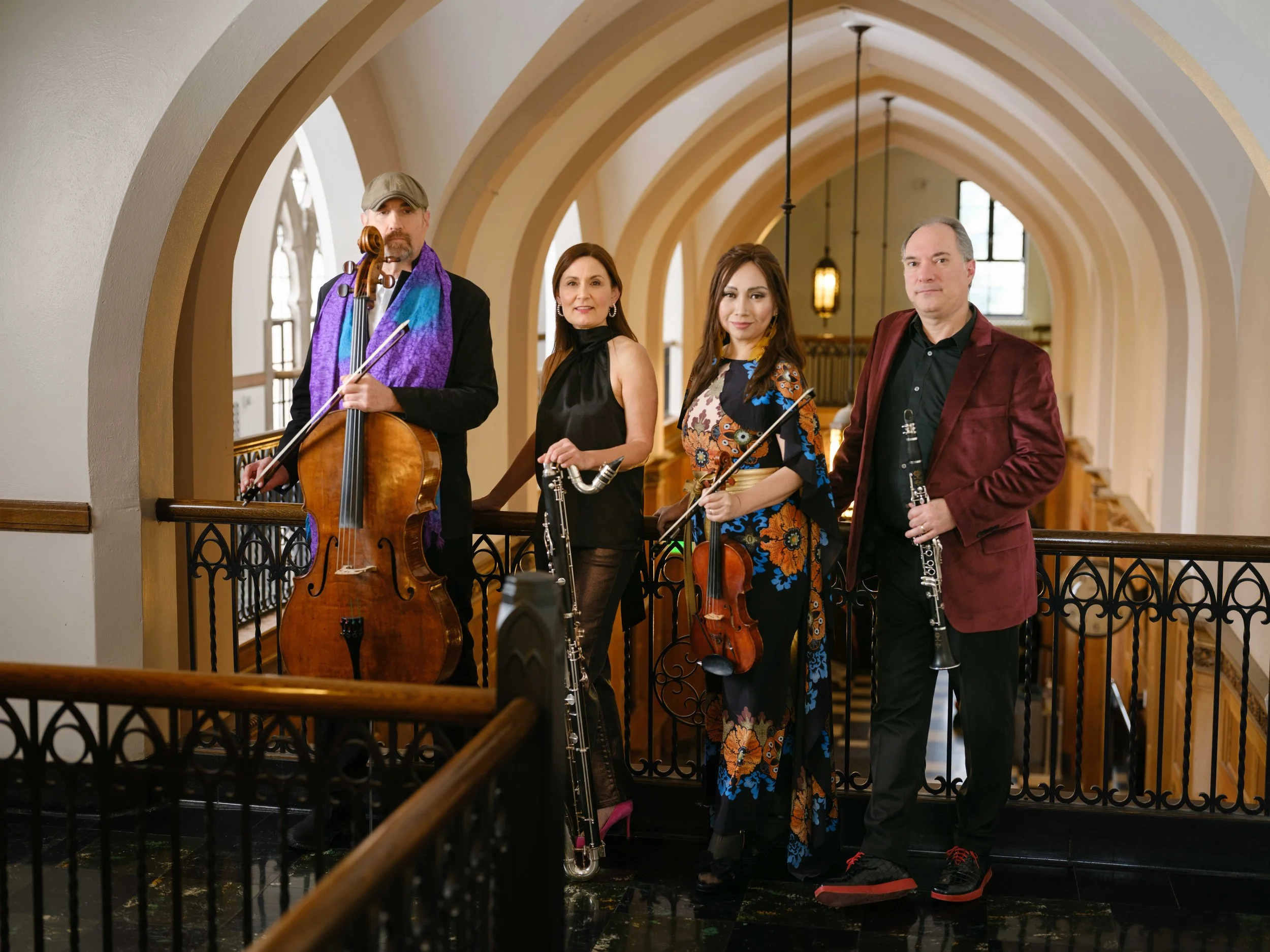 A group of four musicians with string and wind instruments standing on a balcony in a building with arched ceilings and windows.