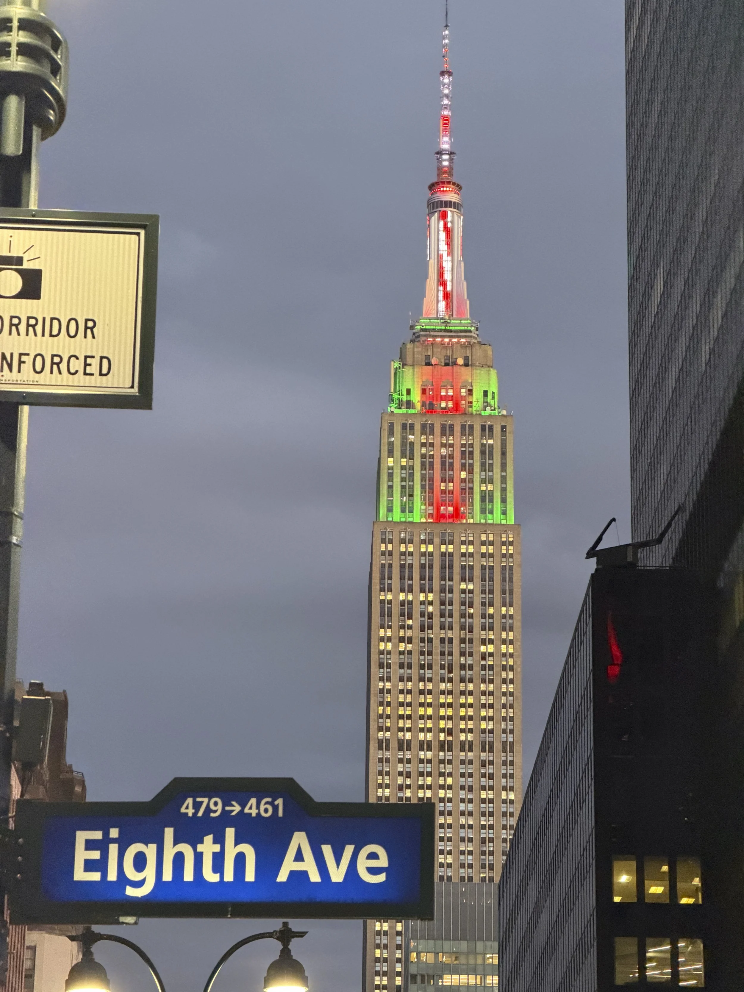 The Empire State Building lit up in red, white, and green lights at night, with street signs for Eighth Avenue and a corridor sign visible in the foreground.
