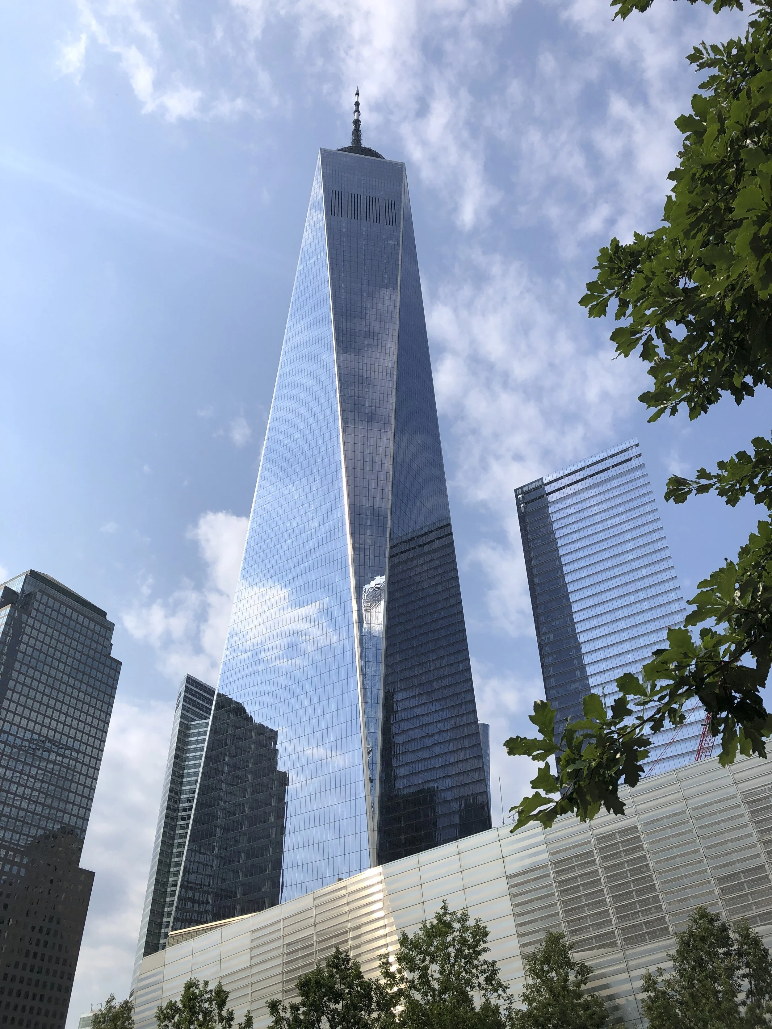 The tall modern skyscraper known as One World, in New York City,  with a glass facade reflecting the sky and surrounding buildings, set against a partly cloudy sky with trees in the foreground.