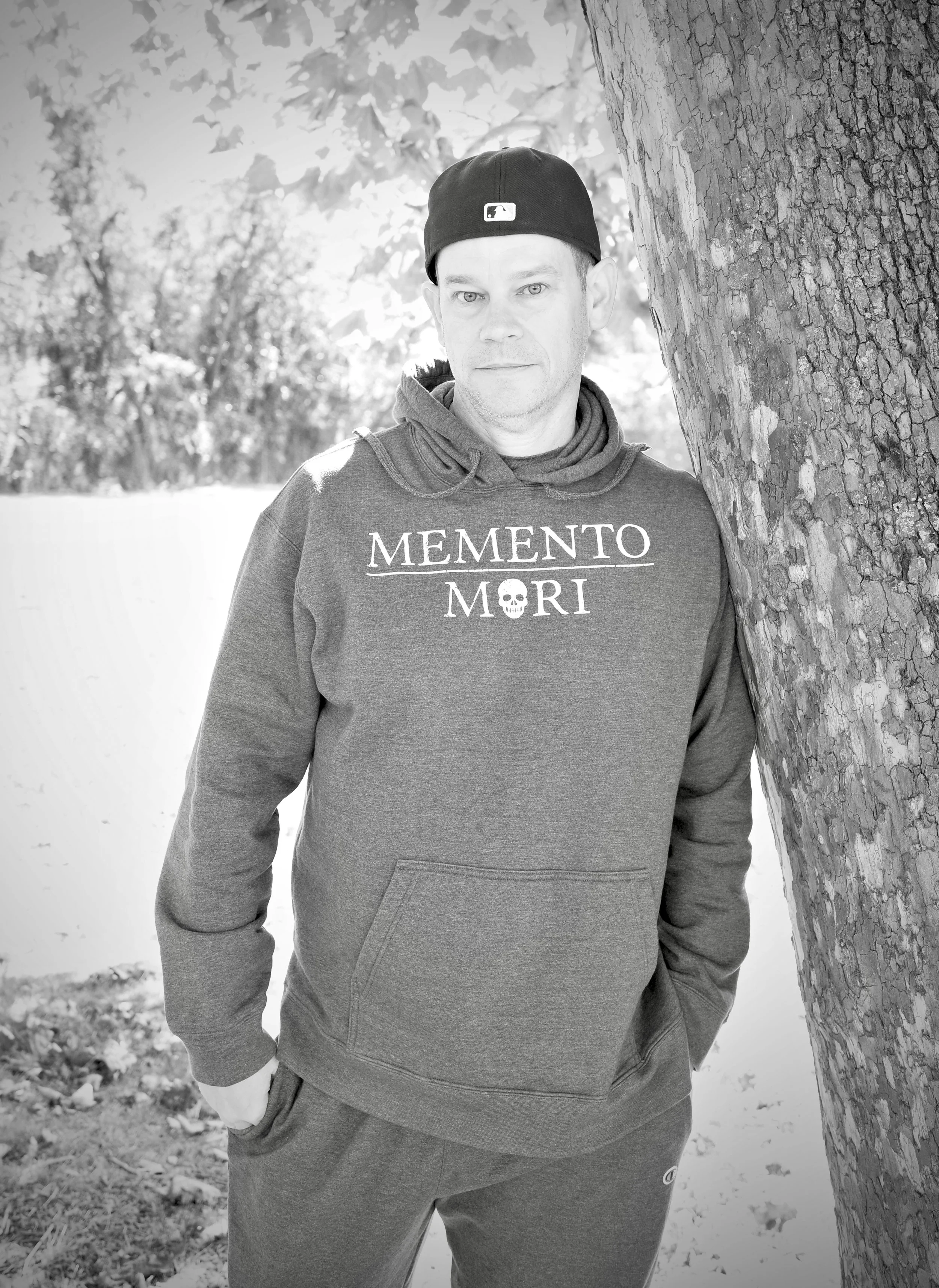 A man wearing a hoodie about Stoicism, Memento Mori,  and a baseball cap, standing outdoors next to a tree with one hand in his pocket, in a black-and-white photo.