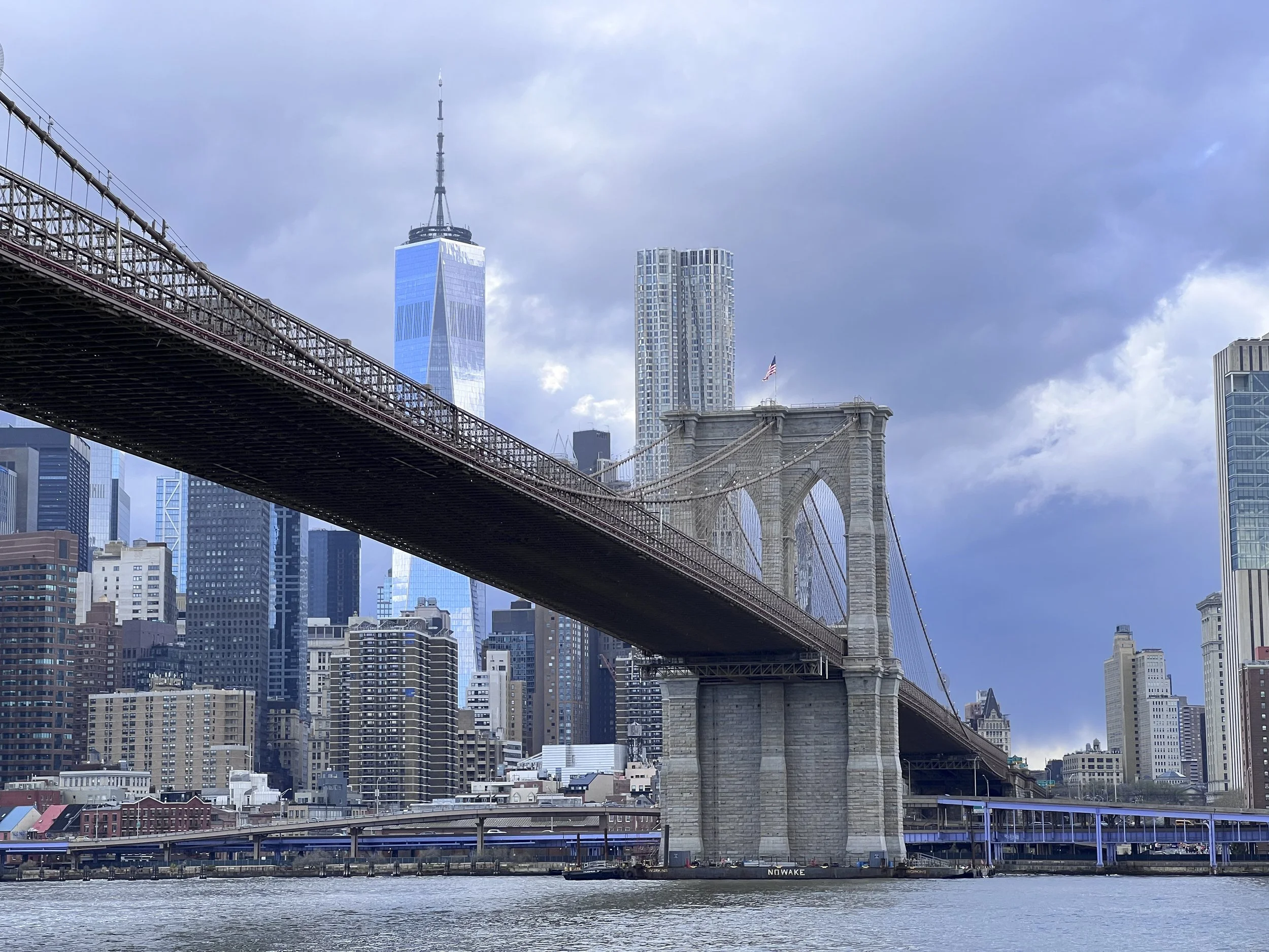View of the Brooklyn Bridge with Manhattan's skyline in the background, including One World Trade Center, under a cloudy sky, over the East River.