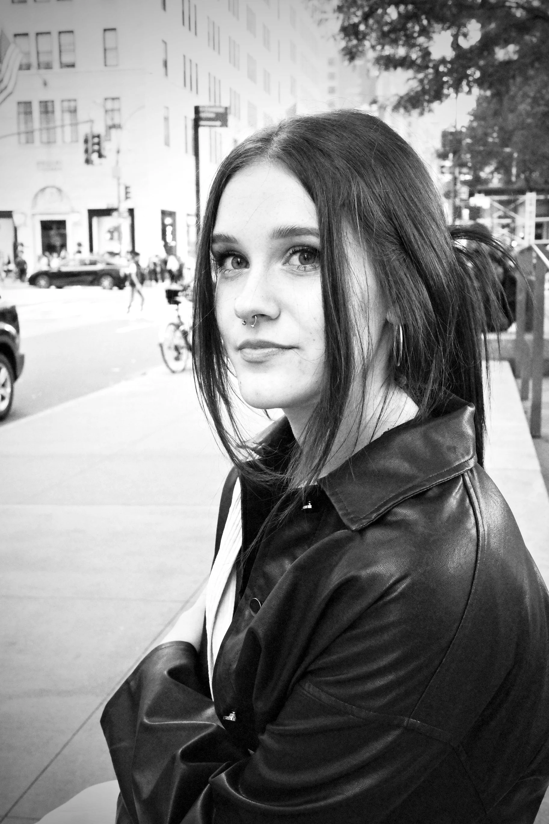 Ken's daughter with straight dark hair and a septum piercing, sitting on a city sidewalk in New York City near 5th Avenue, looking into the camera. She wears a leather jacket, with a busy city street in the background.