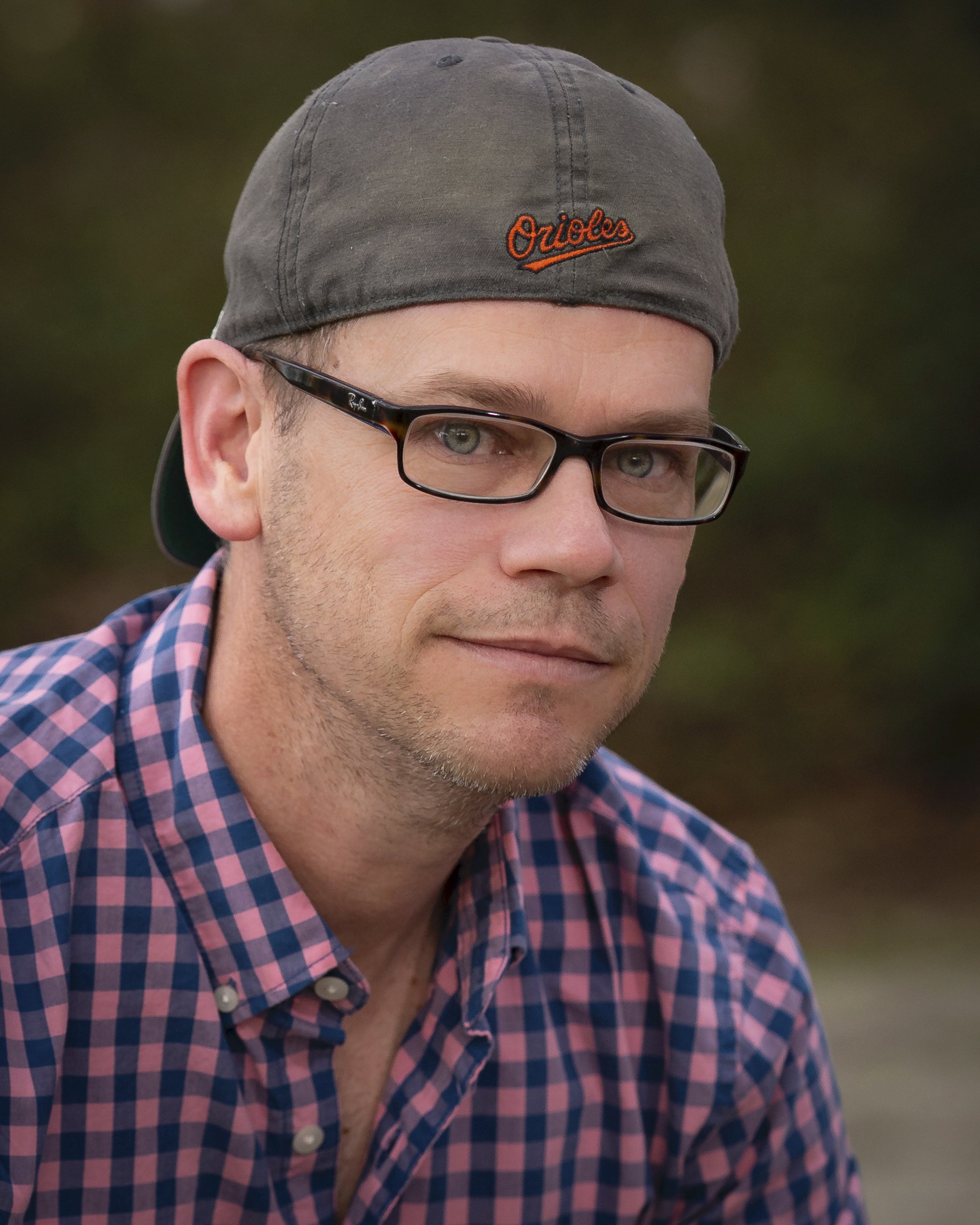 A man wearing glasses, a gray cap with 'Orioles' embroidered on it, and a pink and blue checkered shirt, outdoors with a blurred nature background.