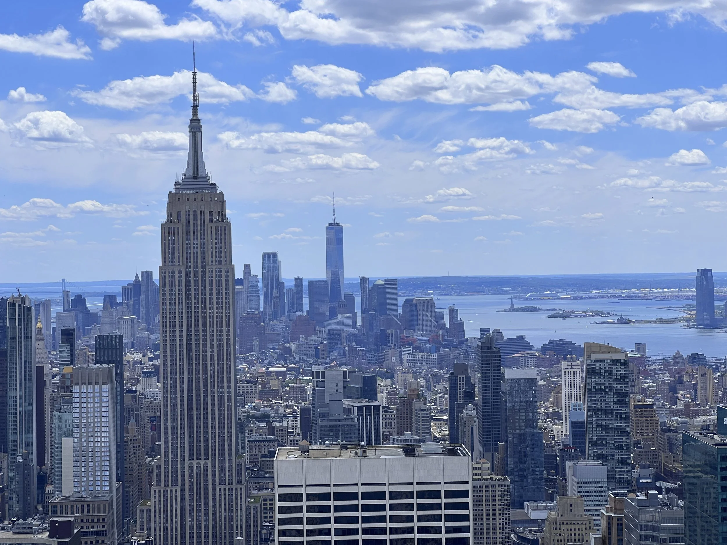 A cityscape featuring the Empire State Building and the One World Trade Center in New York City with a river in the background and a partly cloudy sky.