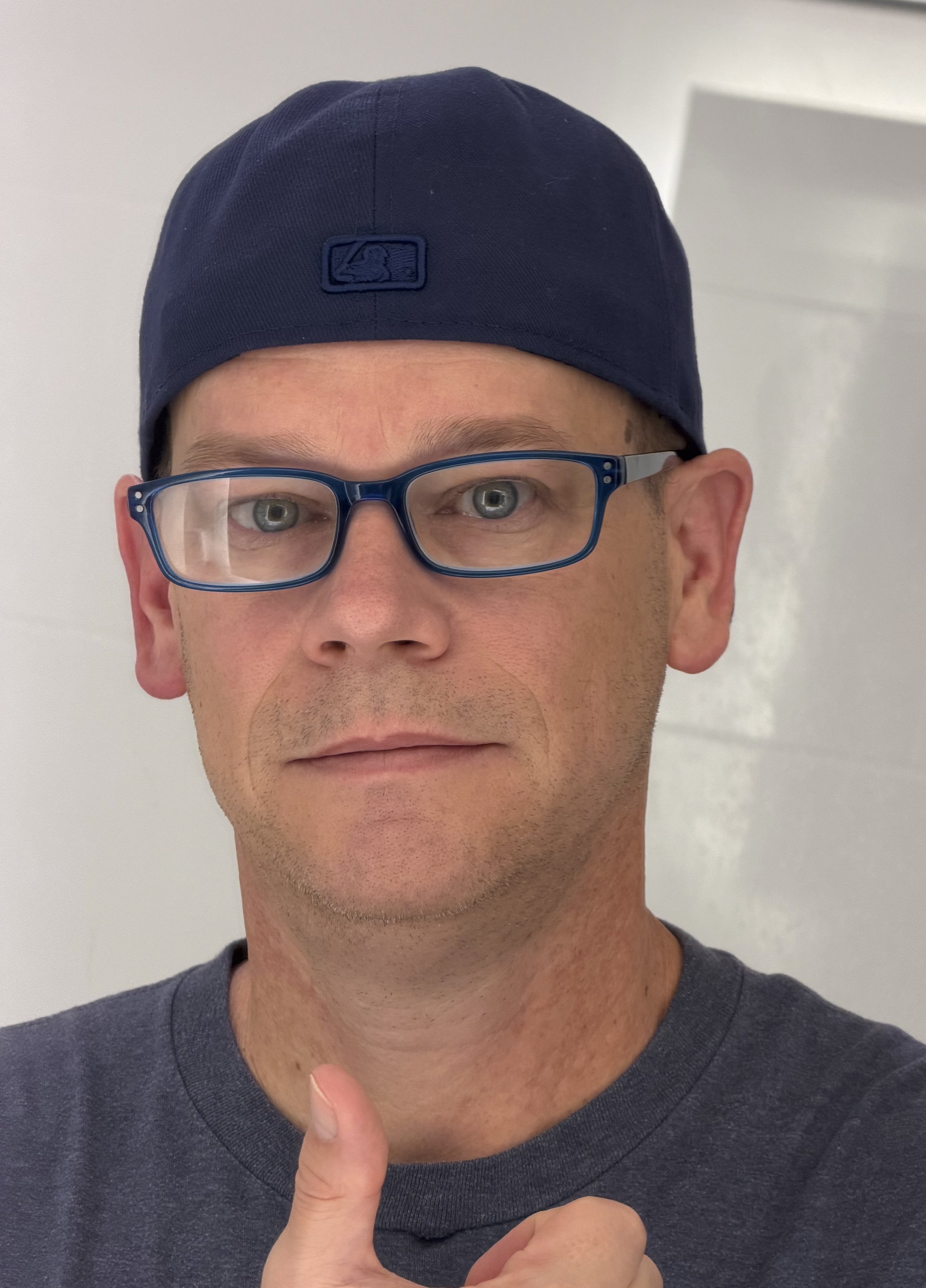 A man wearing a navy blue baseball cap, blue glasses, and a dark gray t-shirt is giving a thumbs-up gesture and looking into the camera.